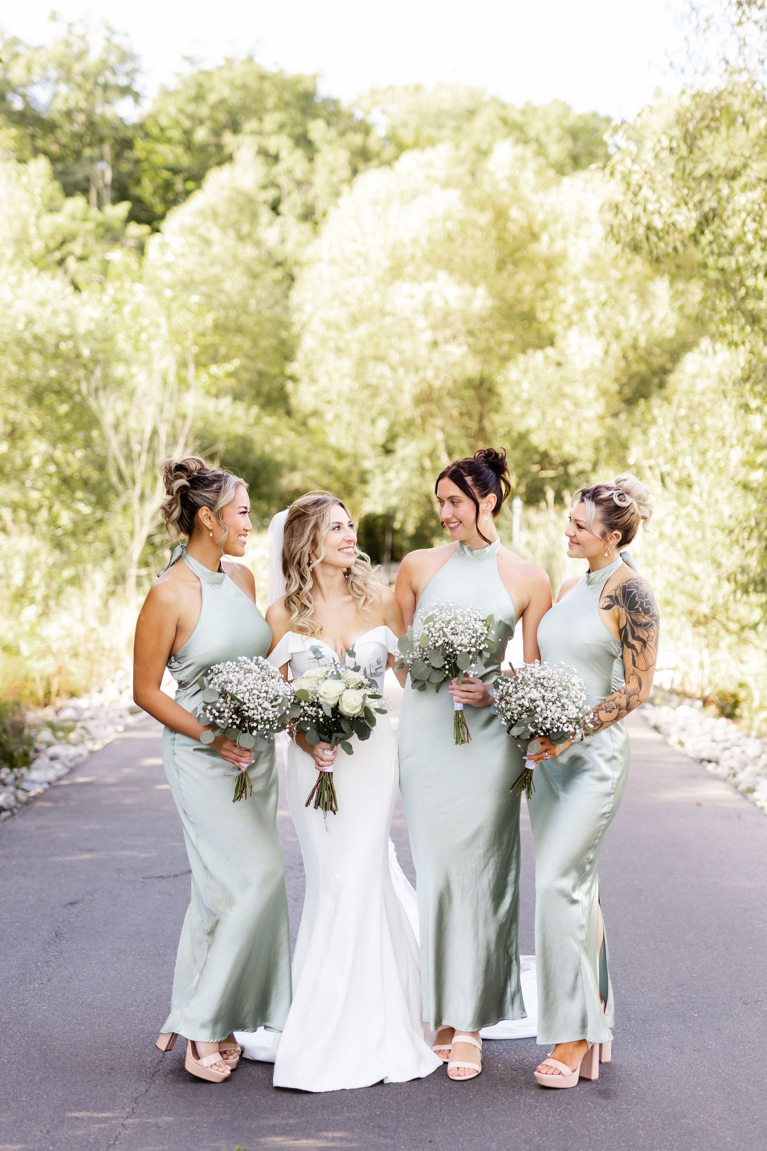 Wedding party in sage green dresses and white gown posing together on paved path.