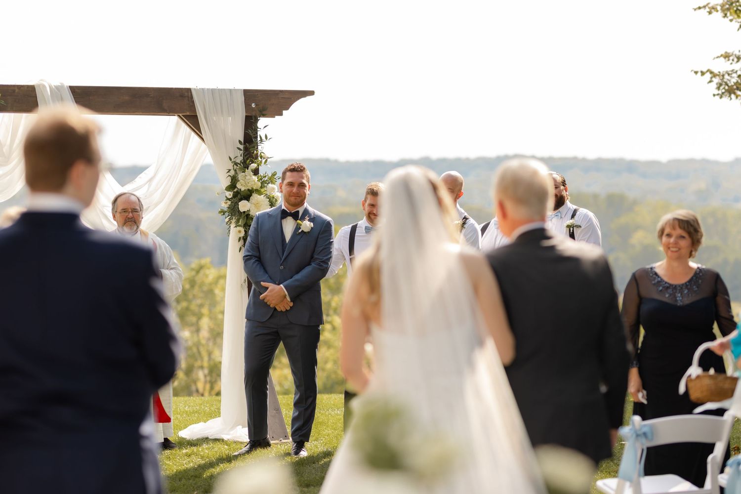 Outdoor wedding ceremony scene with guests seated on white chairs under trees.