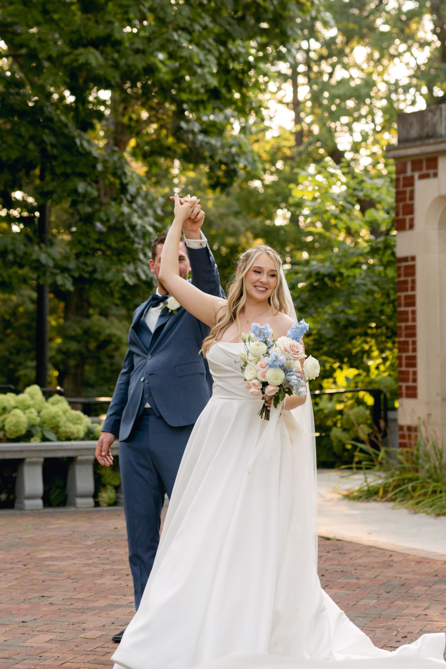 Bride in flowing white gown twirls on brick patio surrounded by greenery.