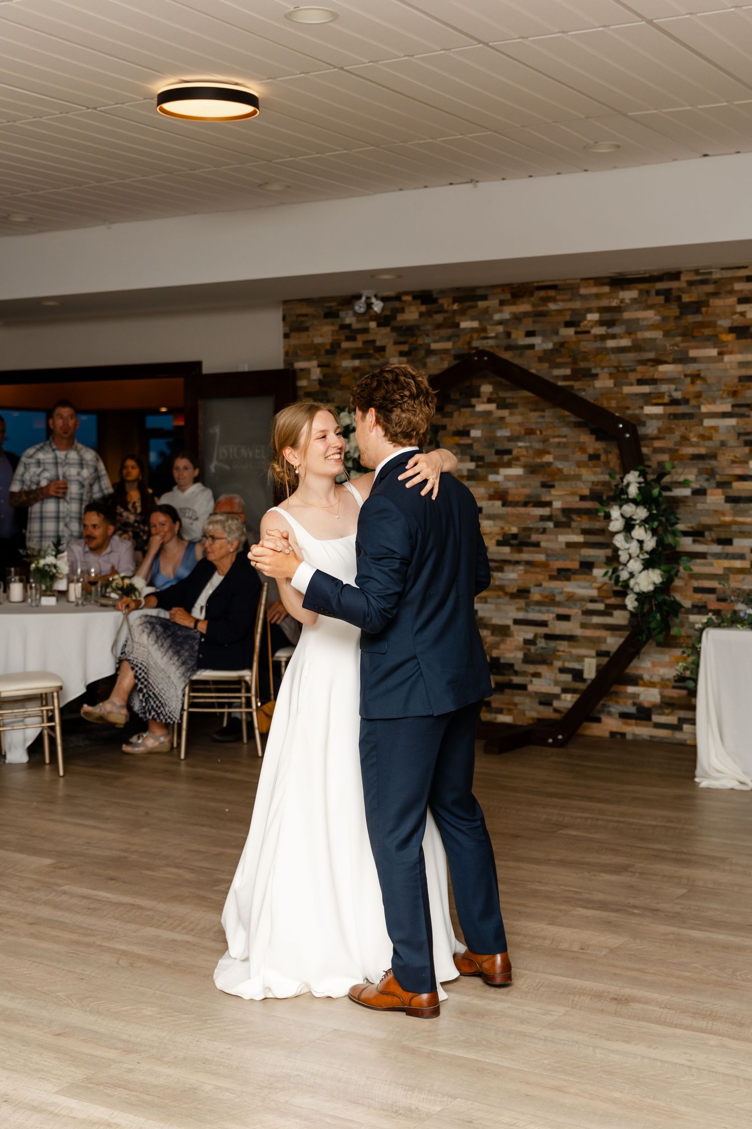 Newlyweds dancing together on wooden floor with stone accent wall in background.