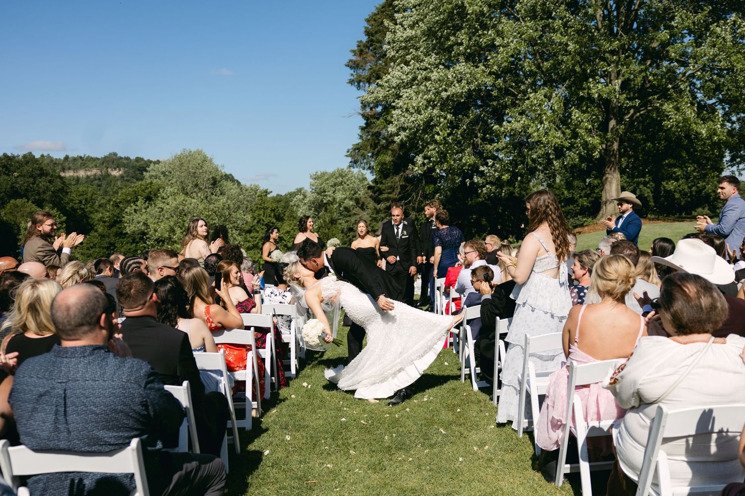 Wide view of outdoor wedding ceremony with guests seated on a green hillside.