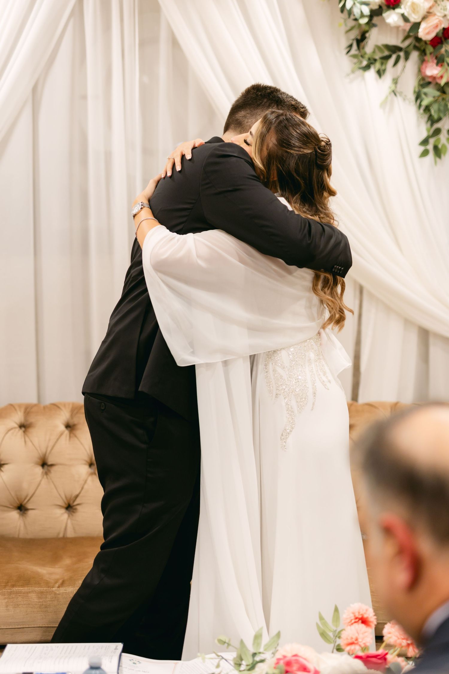 A couple embraces during their wedding ceremony with elegant white drapes and flowers in the background.