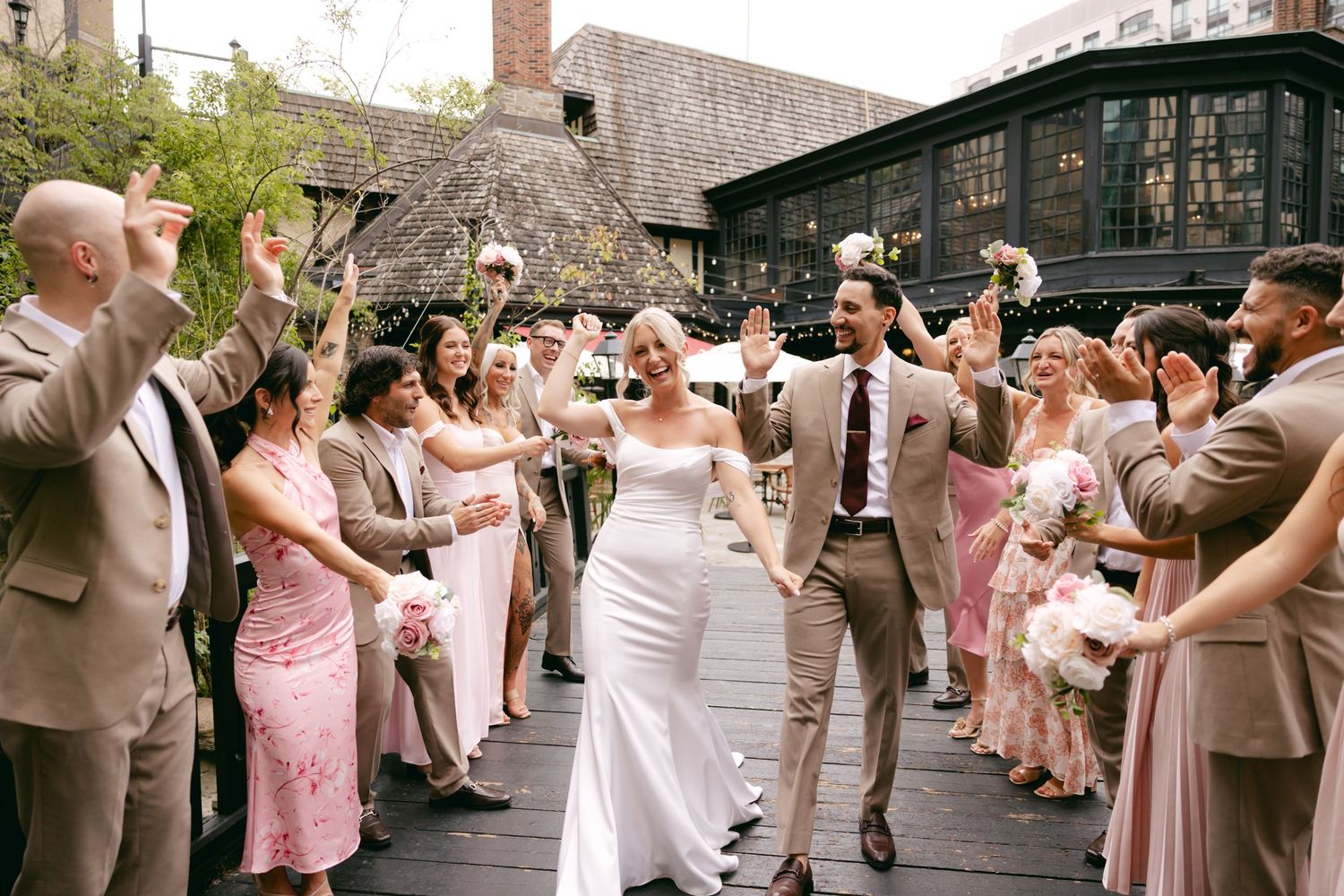 Wedding party celebrates with joyful dancing in an outdoor venue with pink and tan attire.