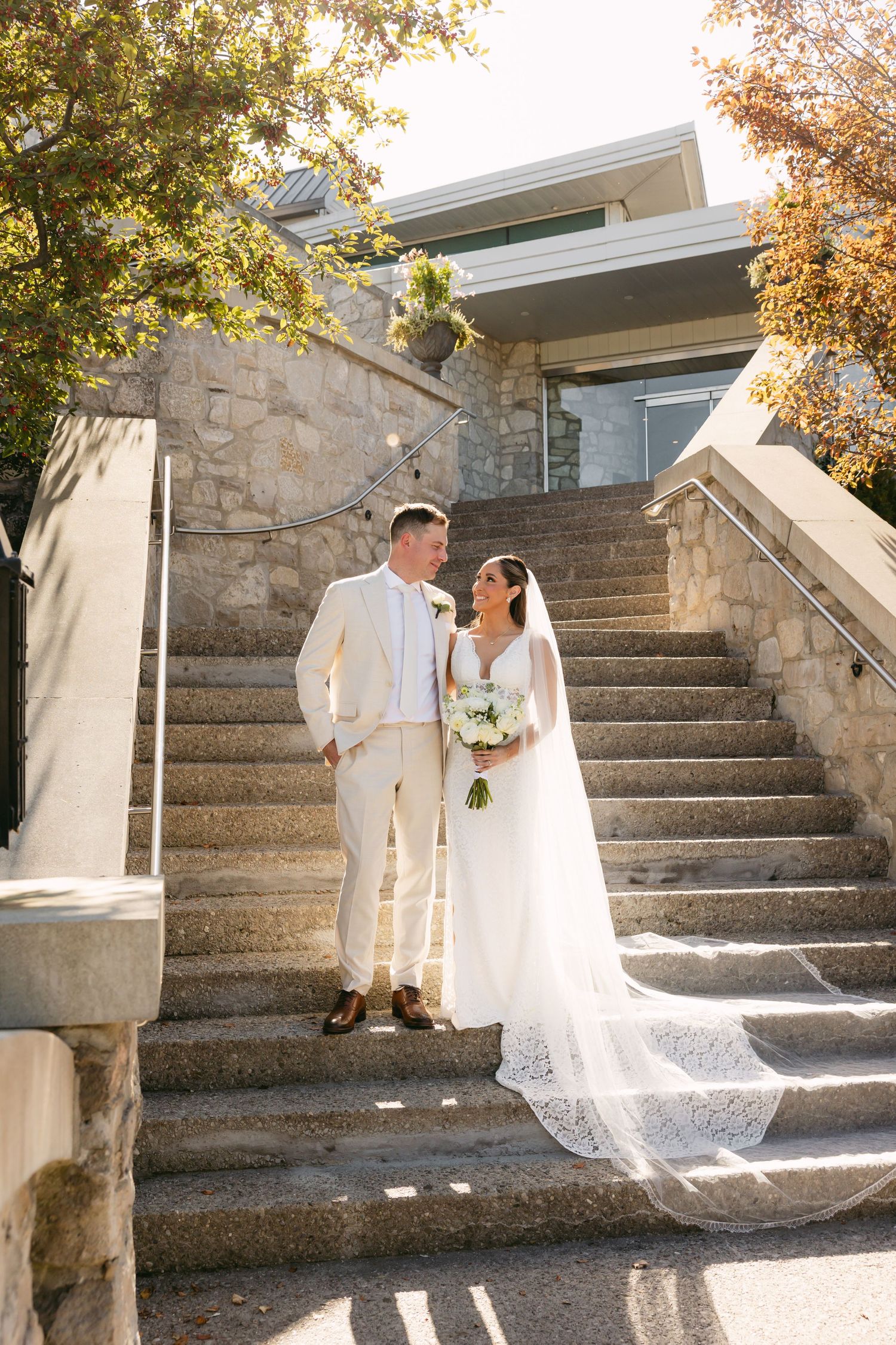 Sunlit wedding photos on stone steps showcase a couple in white attire against autumn foliage.