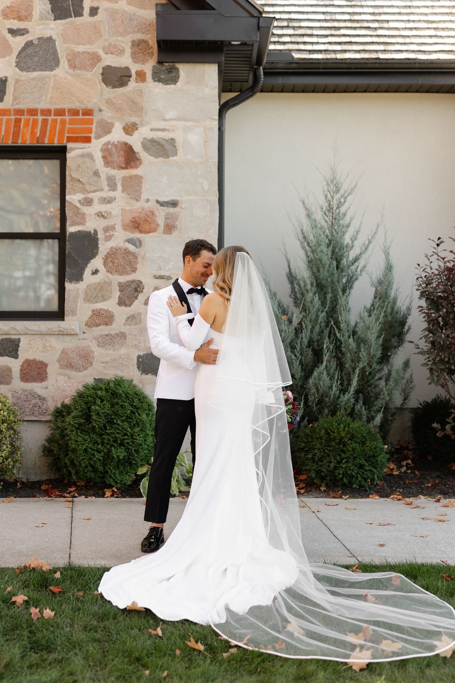 Wedding couple poses outside stone and white building with long flowing veil.