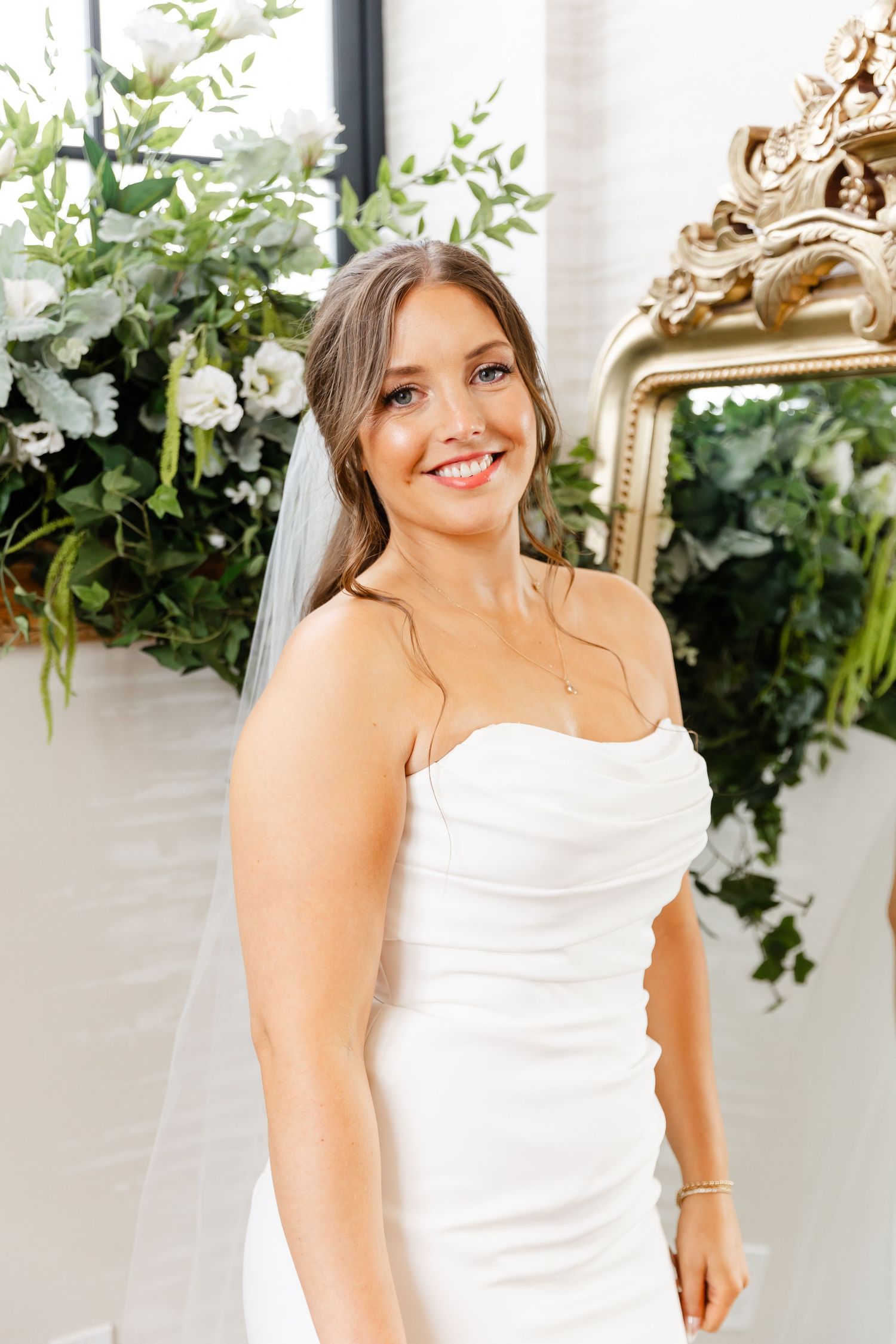 Radiant bride in a strapless white gown smiles beside lush floral arrangements and a gold ornate mirror.
