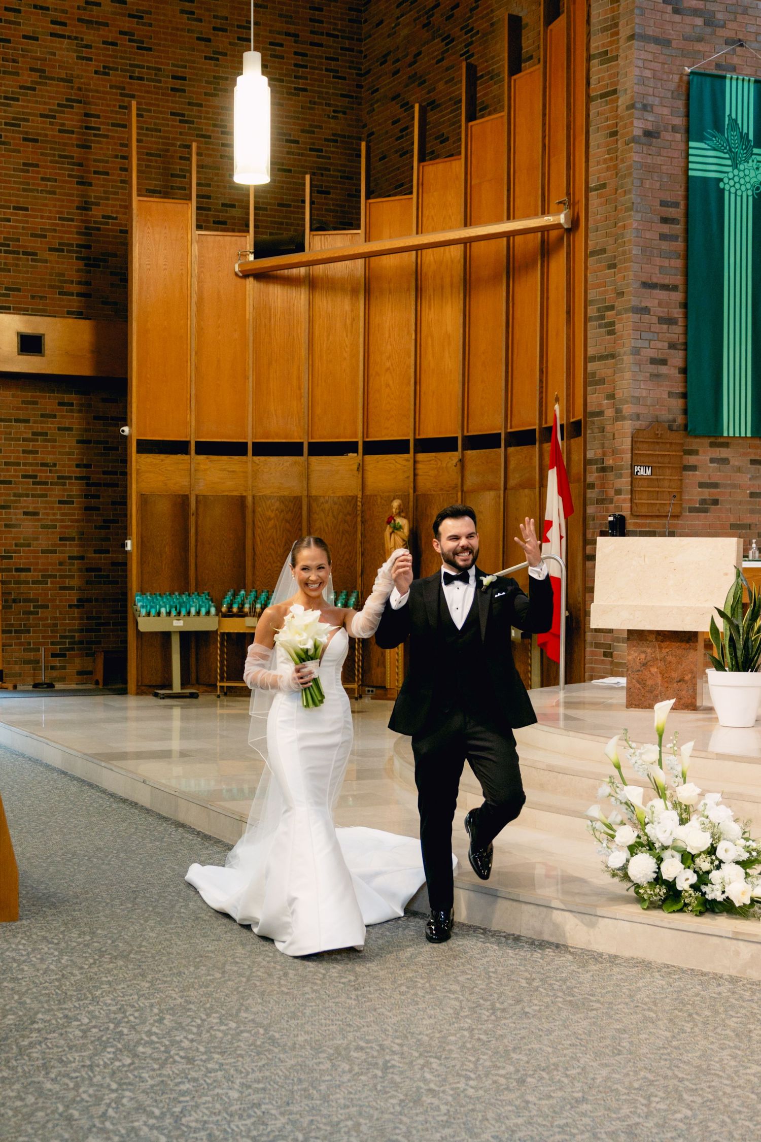 Joyful newlyweds celebrate walking back down the aisle in a modern church, bride in white gown, groom in black tuxedo.