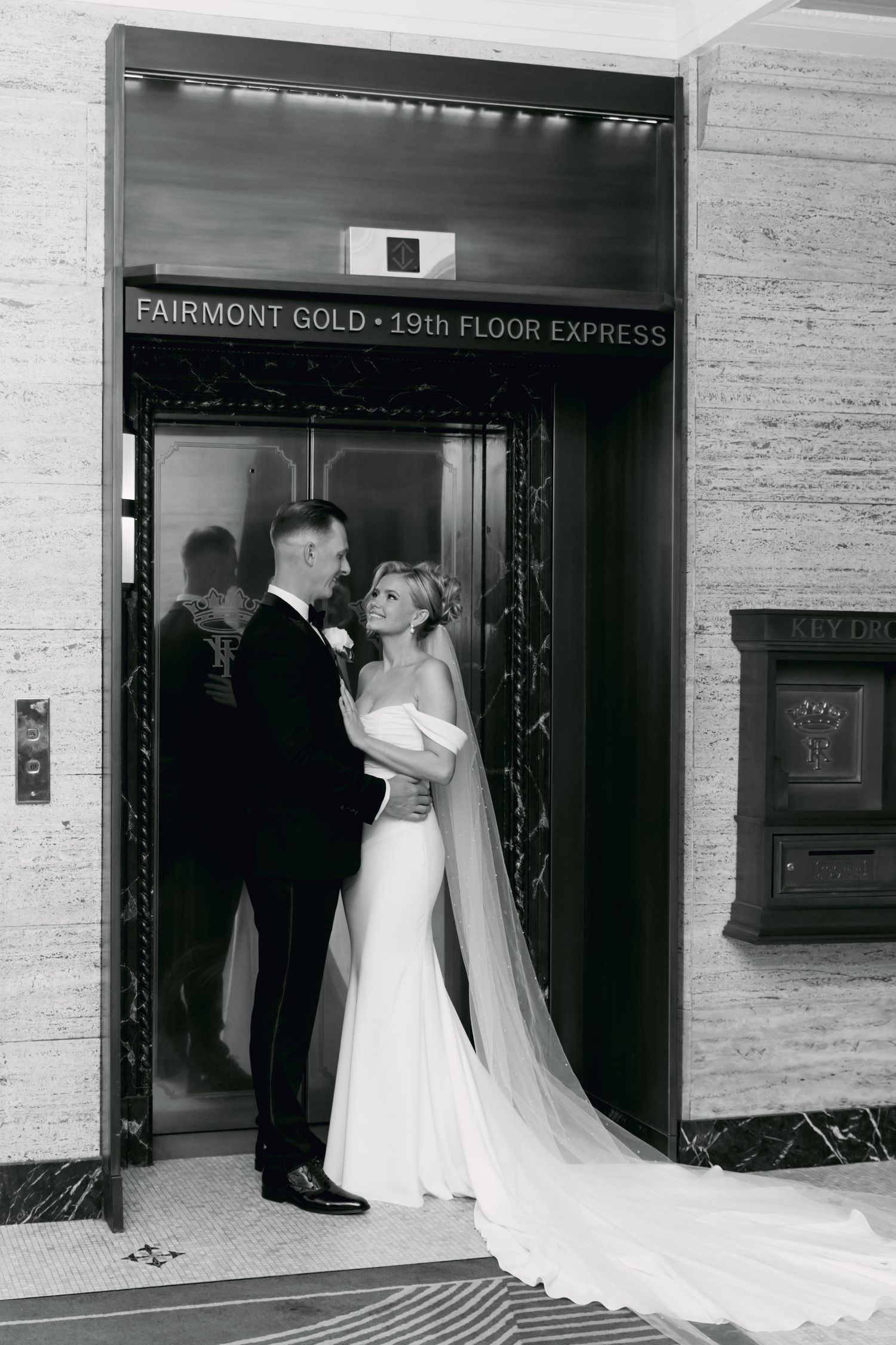 Bride and groom embracing in front of Fairmont Gold 19th Floor Express elevator in black and white wedding photo.