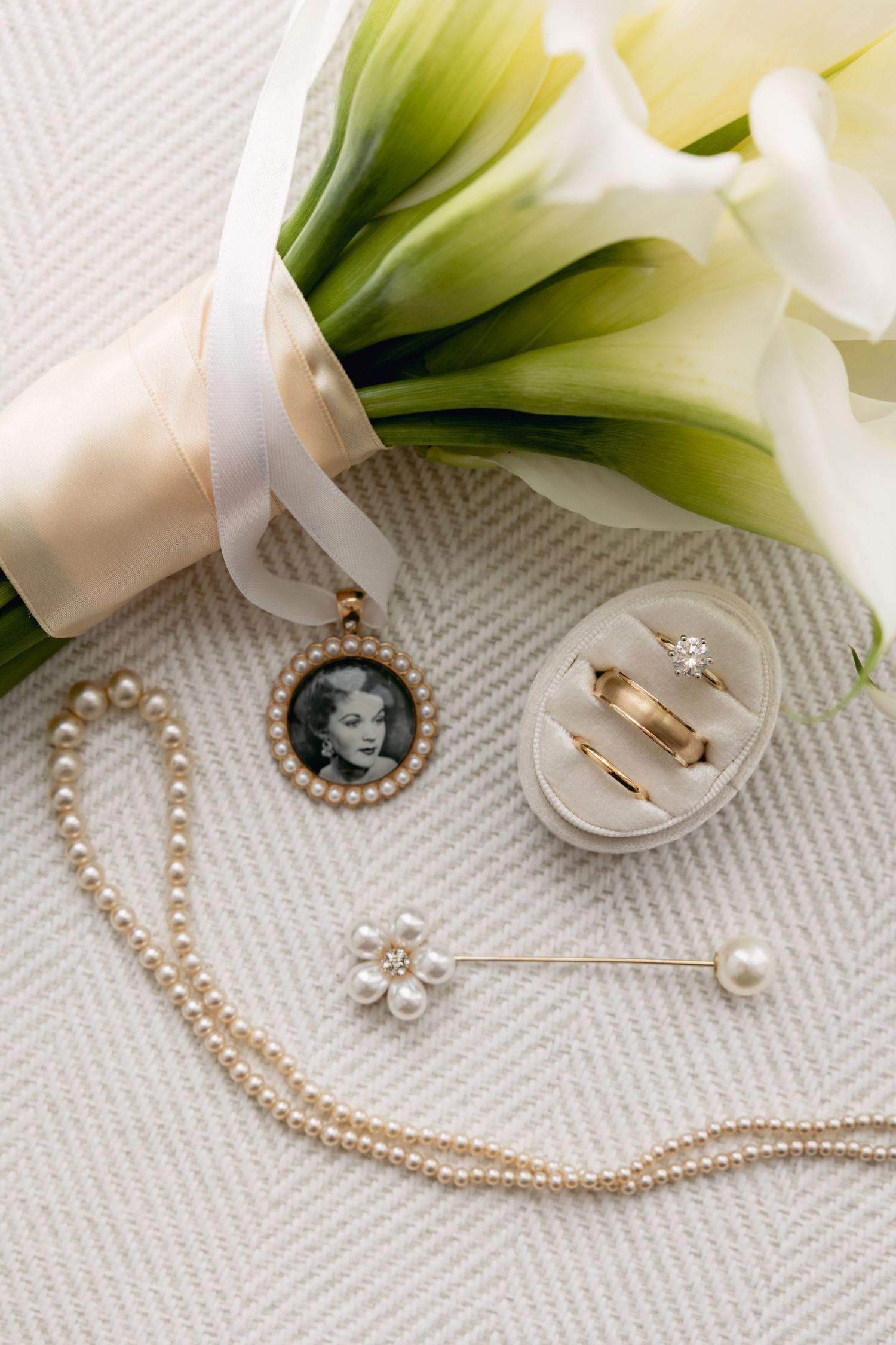 Elegant bridal flat lay with pearl necklace, vintage locket, gold rings, and white calla lily bouquet.
