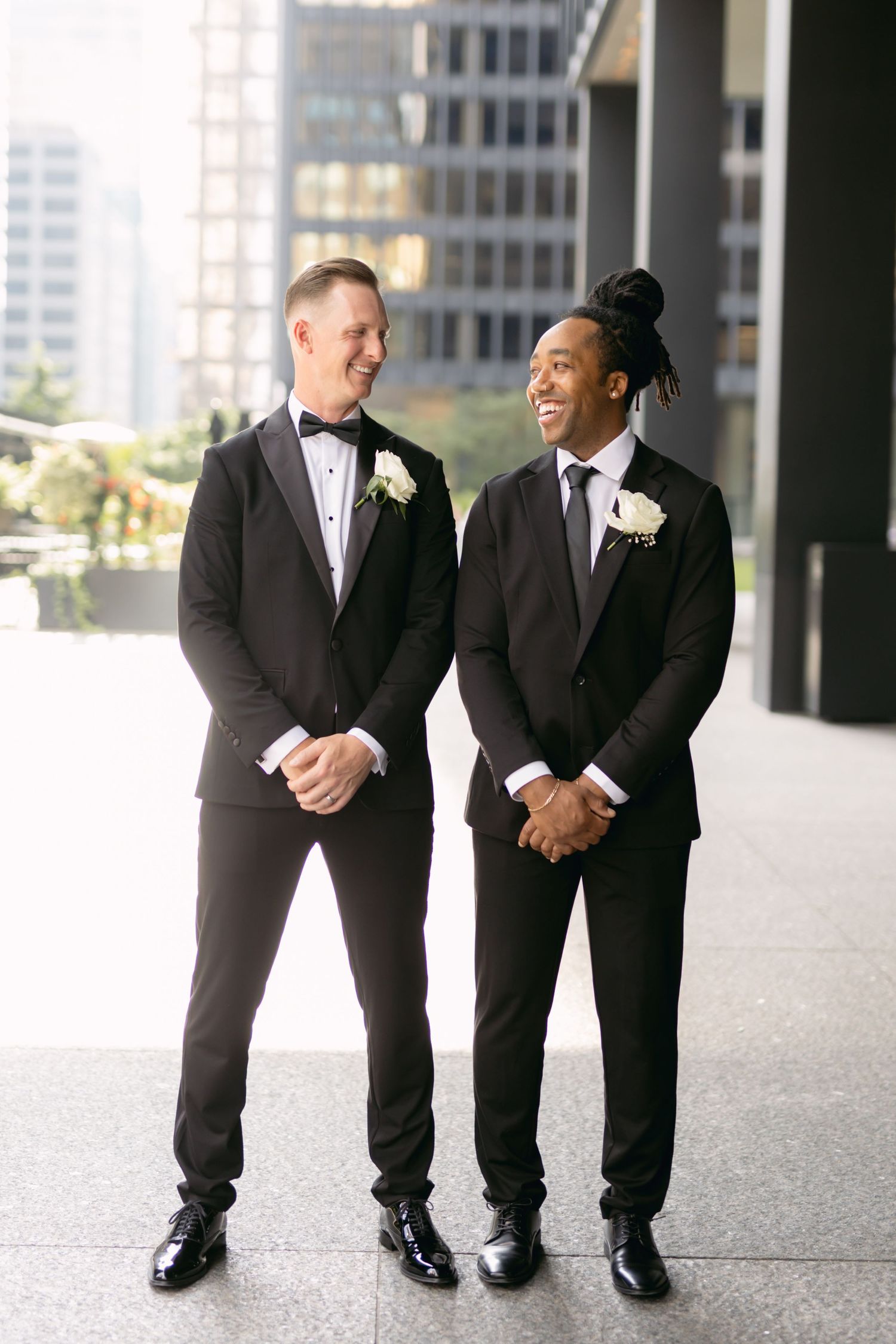 Groom and best man in black tuxedos laugh together outdoors with city skyline in background.