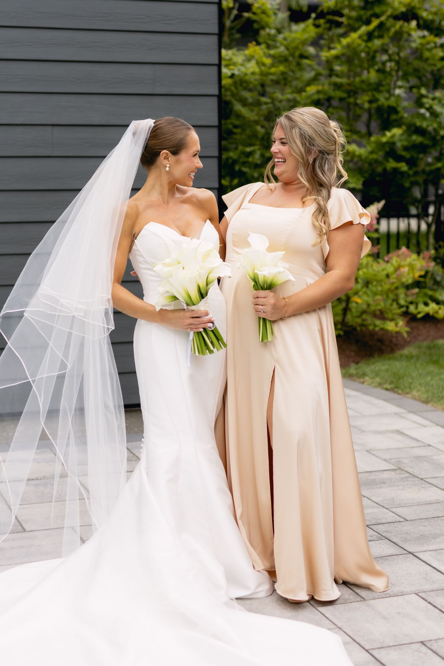 Bride in strapless white gown with veil and bridesmaid in champagne dress hold white calla lily bouquets outdoors.