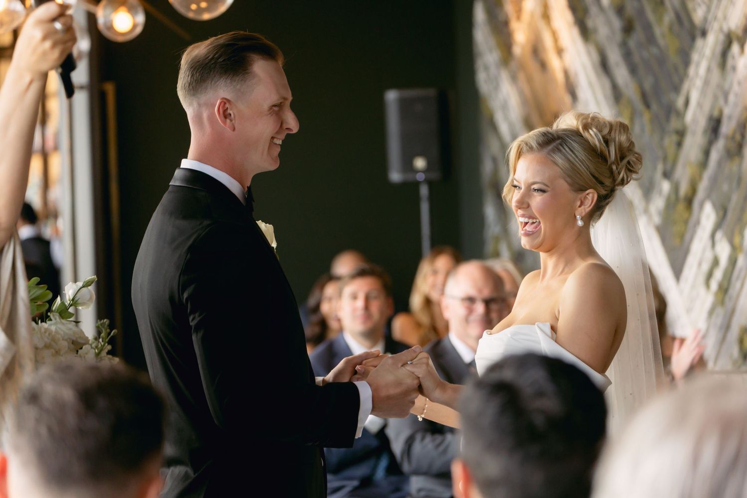 Smiling bride and groom exchanging vows at indoor wedding ceremony with guests seated in background.