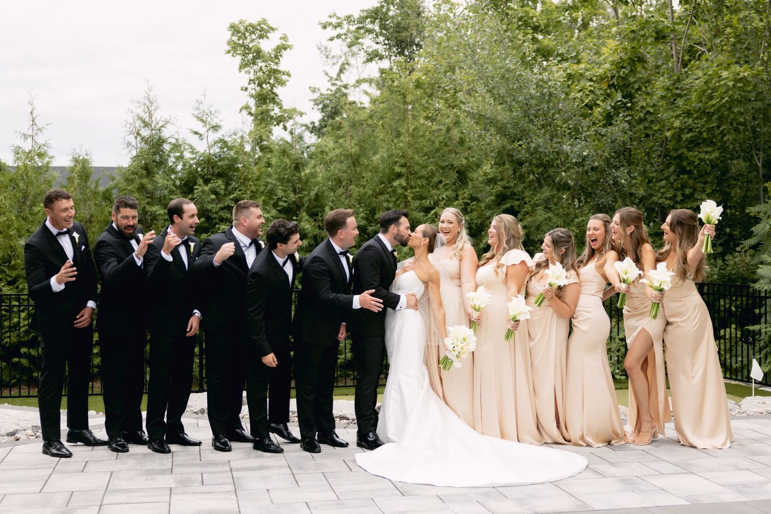 Wedding party with groomsmen in black tuxedos and bridesmaids in champagne gowns posing outdoors.