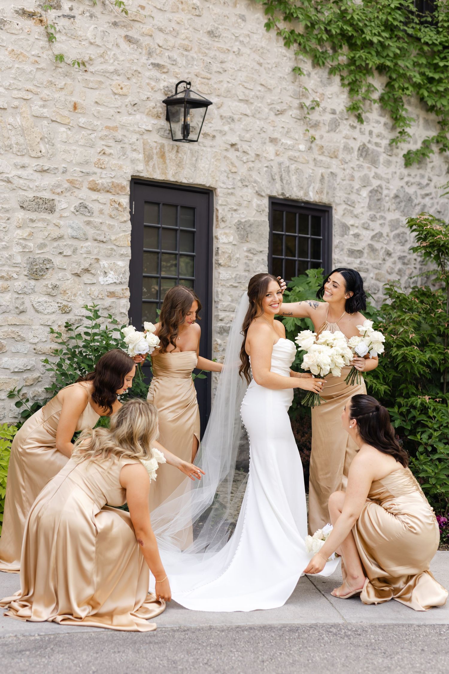 Bride in white gown surrounded by bridesmaids in gold satin dresses adjusting her veil outside stone building.