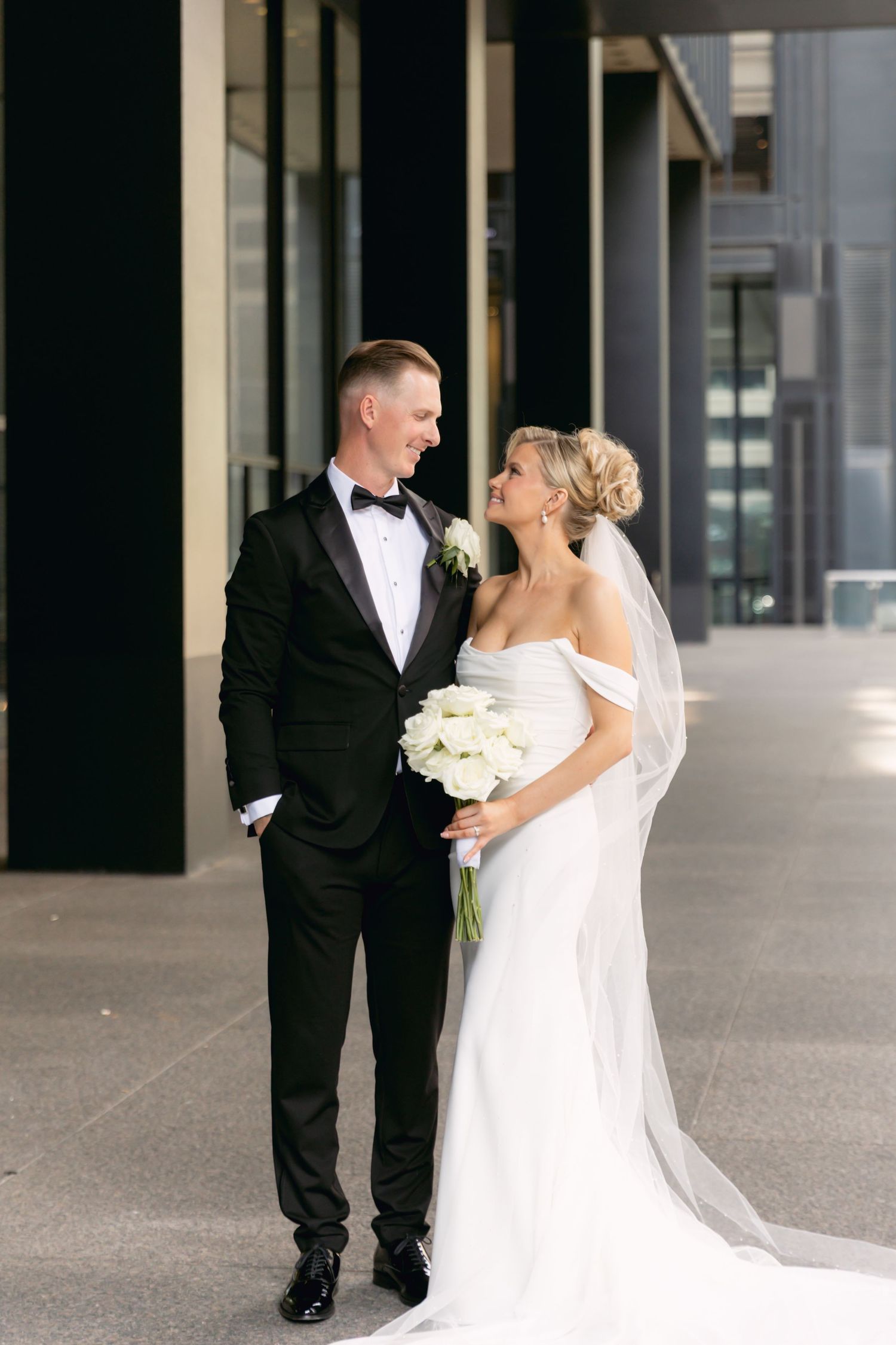 Bride and groom share a romantic moment outdoors, bride holding white bouquet, groom in black tuxedo with bow tie.