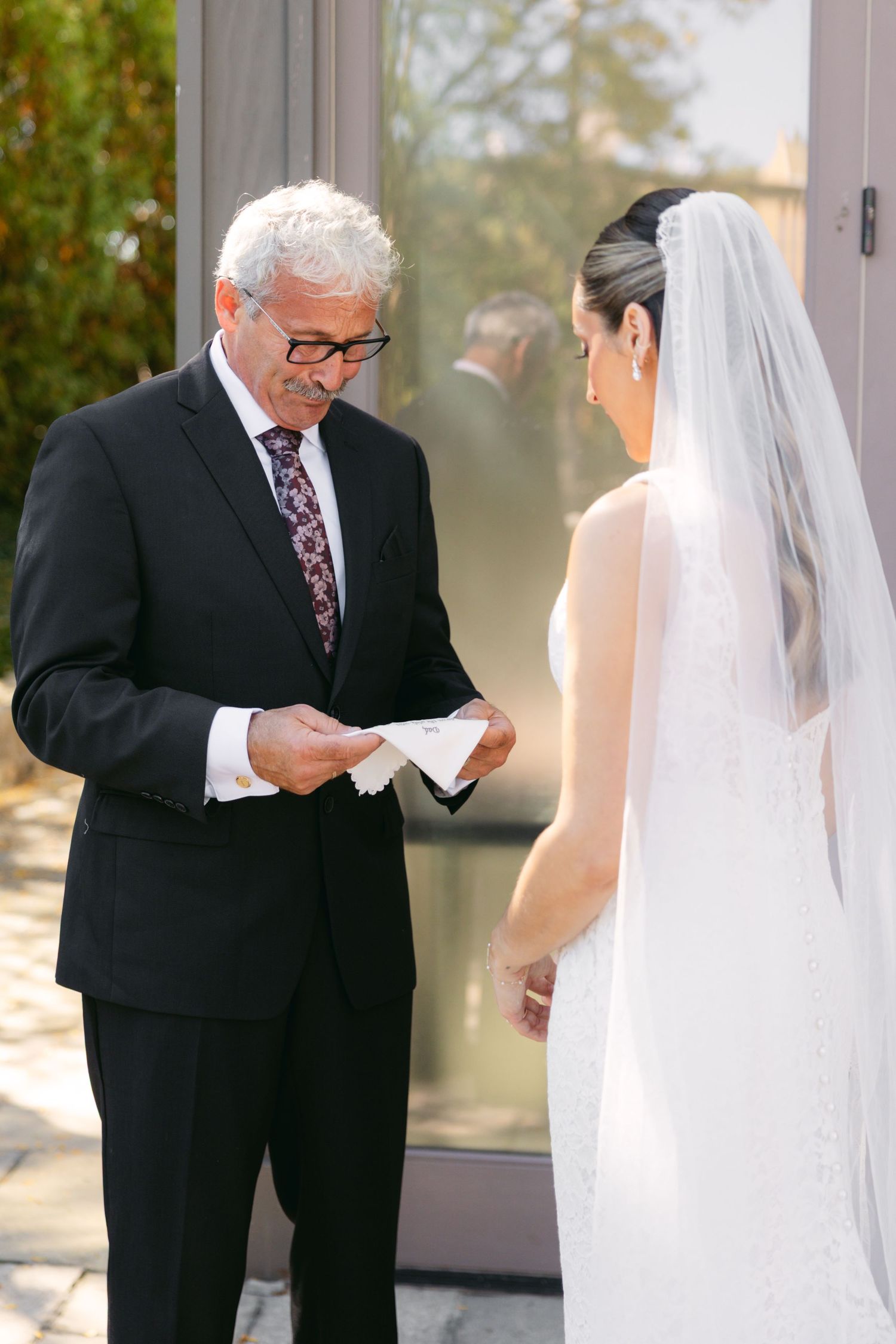 A bride in a white wedding dress receives a reading from an officiant in a black suit during an outdoor ceremony.