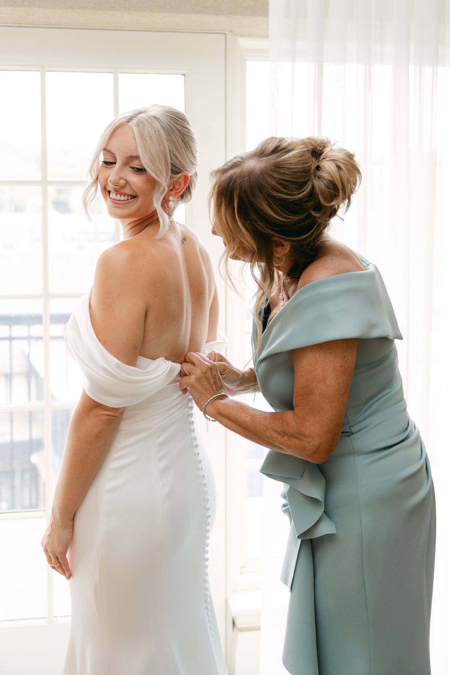 Emotional moment between women in white and sage green dresses during wedding preparations.