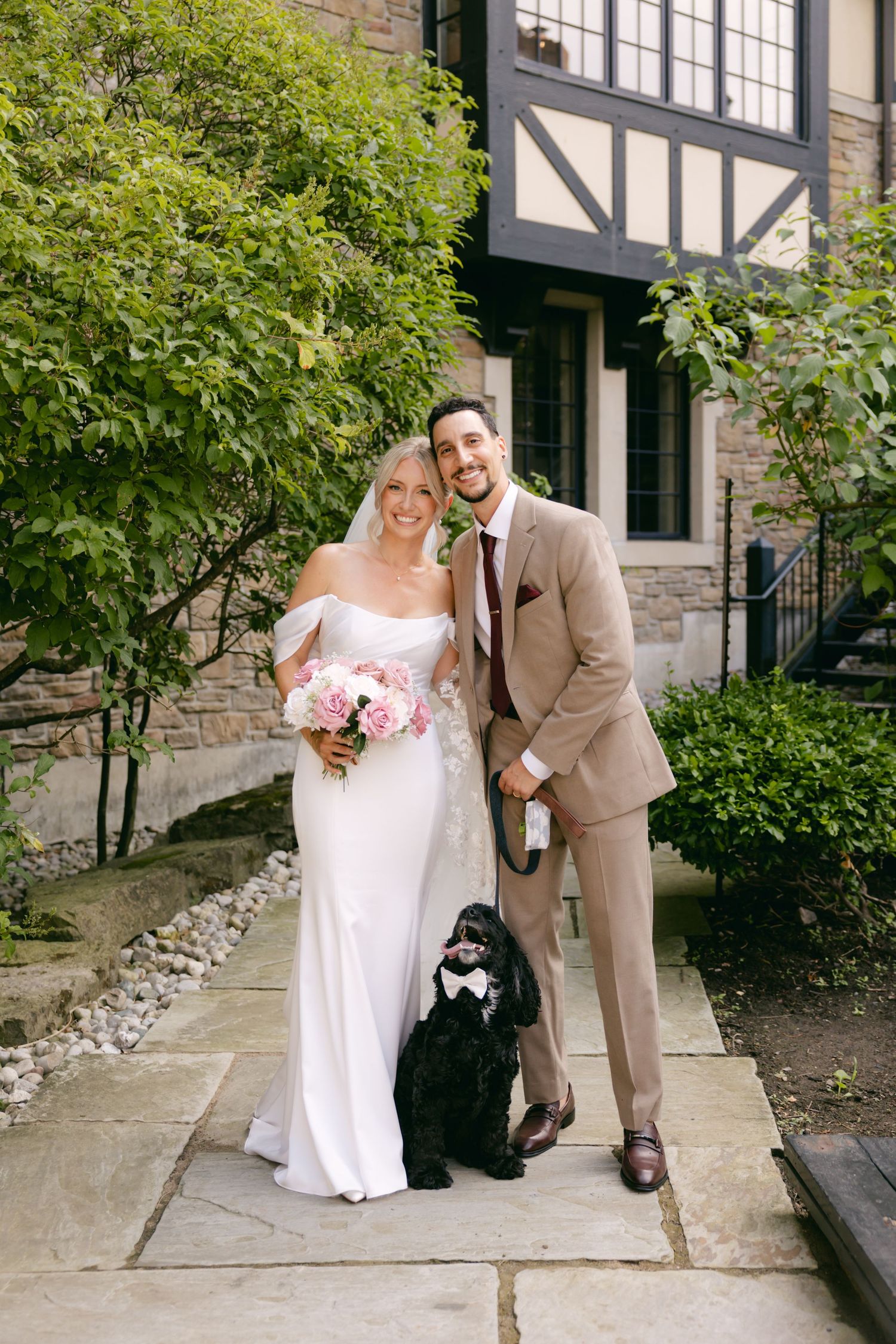 A wedding couple poses with their black dog on stone steps outside a building with green foliage.