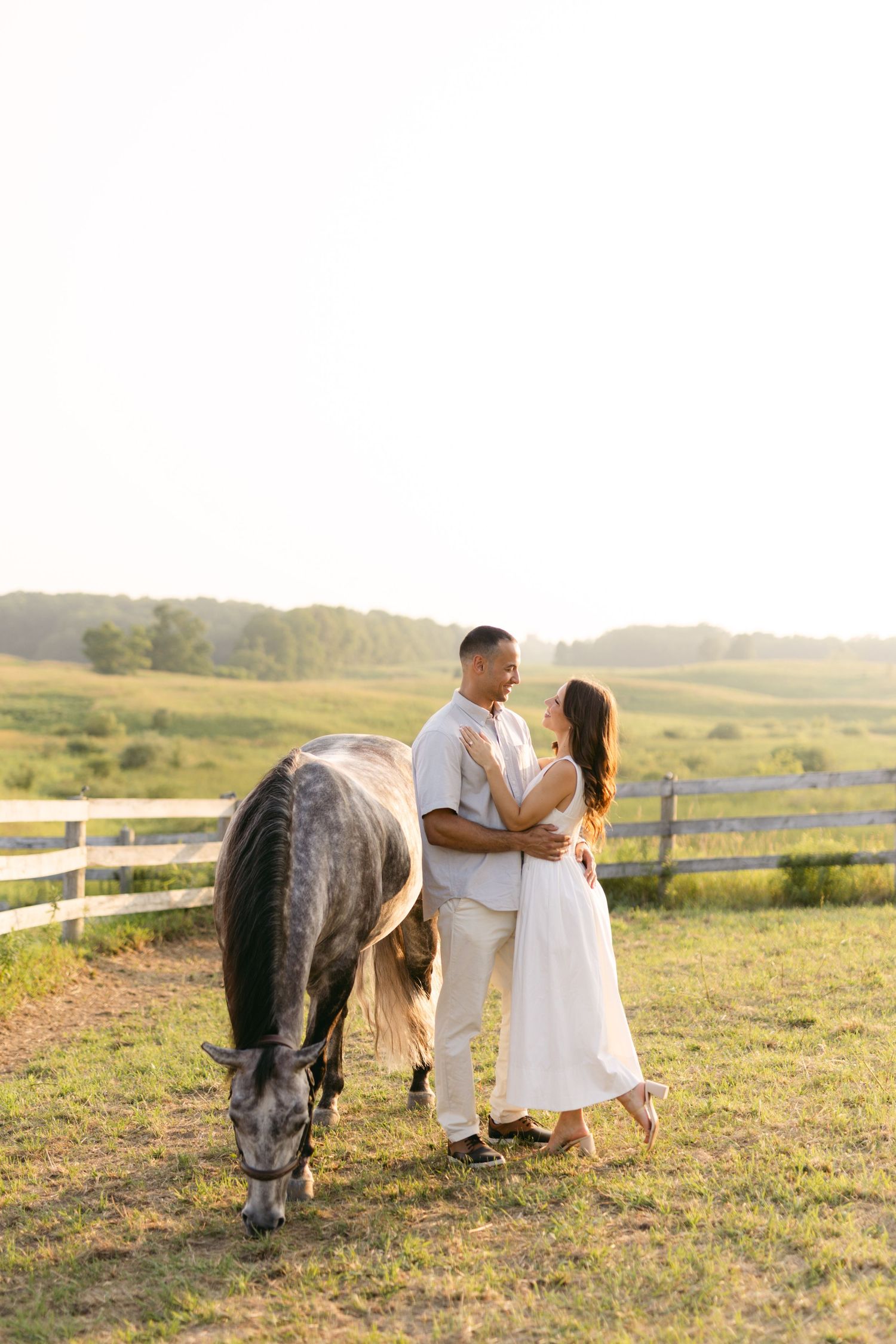 Couple sharing a tender moment in a field at sunset with a horse grazing nearby.