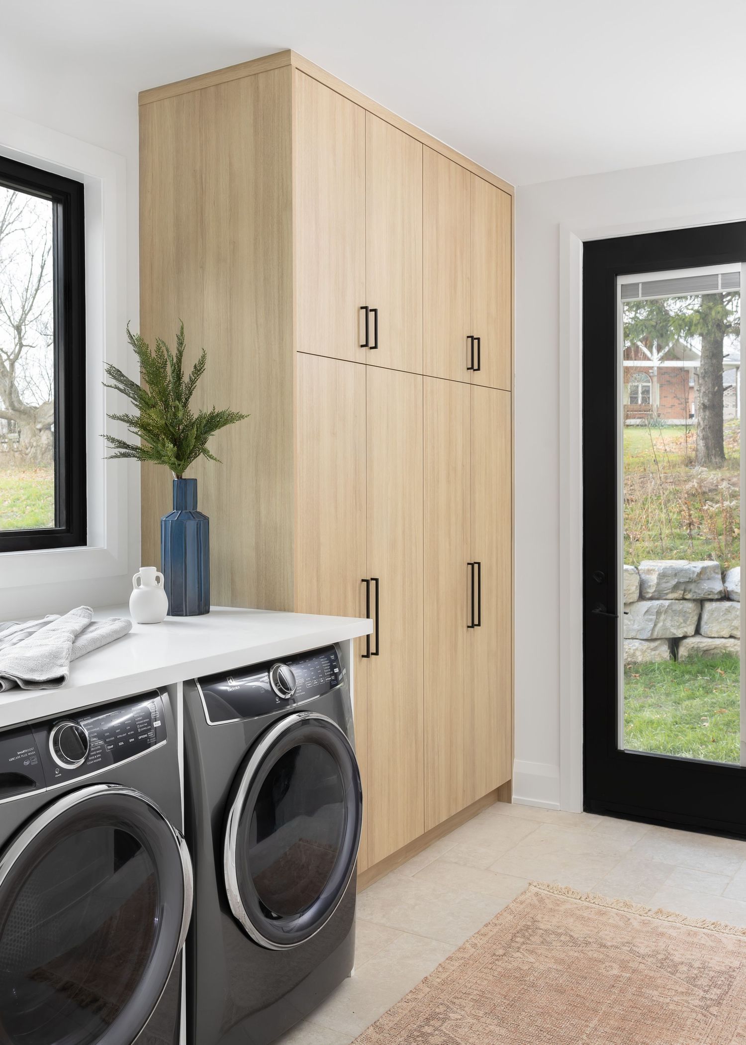 Light wood laundry room cabinets with gray washer and dryer set and black-framed glass door.