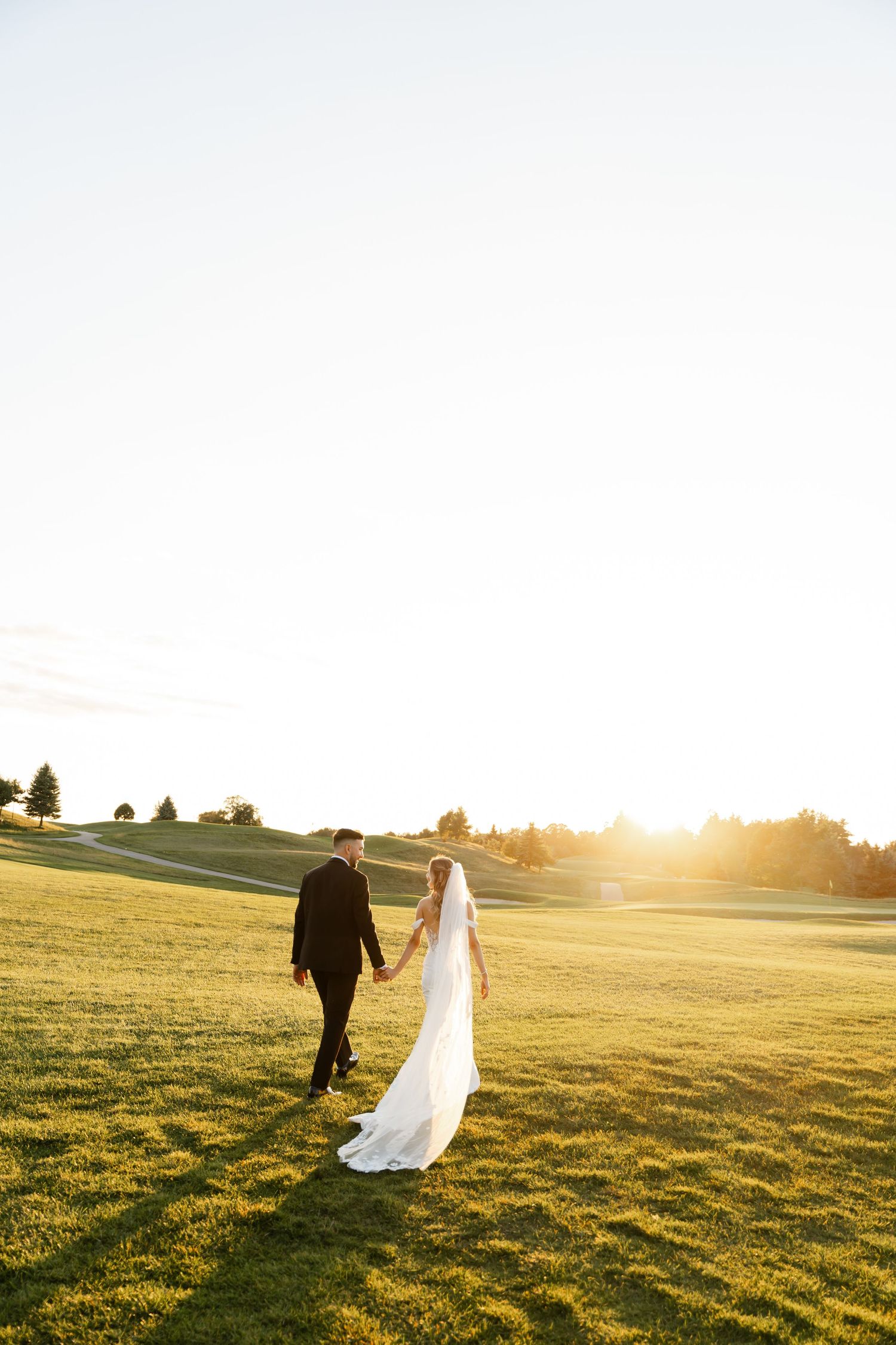 Couple walks across a sunlit golf course during their wedding sunset photo session.