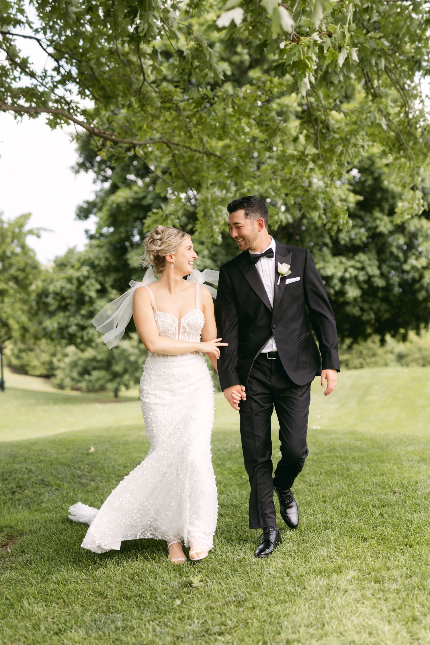 A couple in wedding attire strolling together on manicured grass with trees in the background.