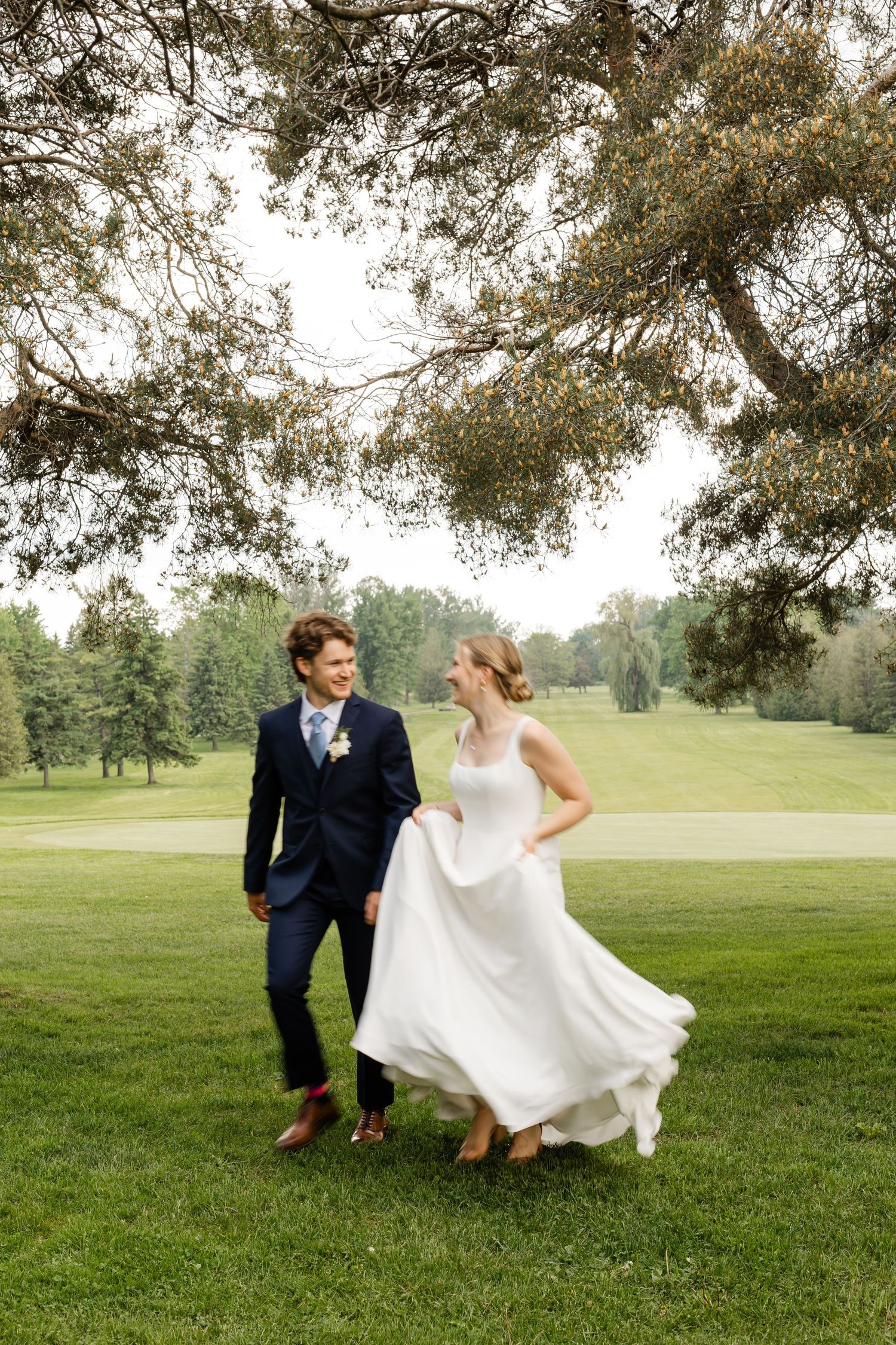 A playful wedding couple dances together on a grassy field under overhanging tree branches.
