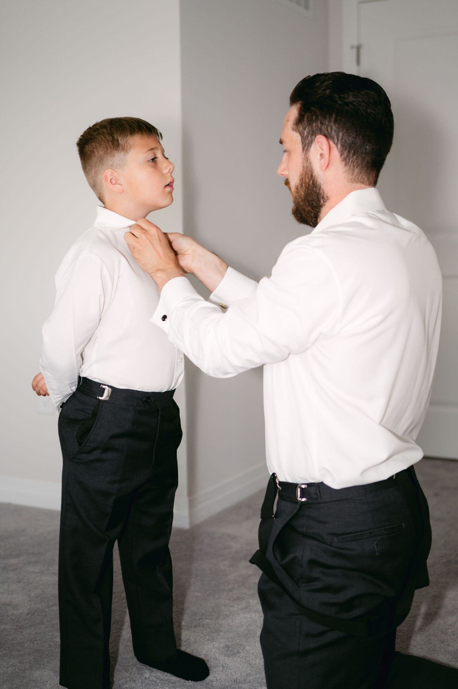 Two people in formal attire interact playfully against a white wall.