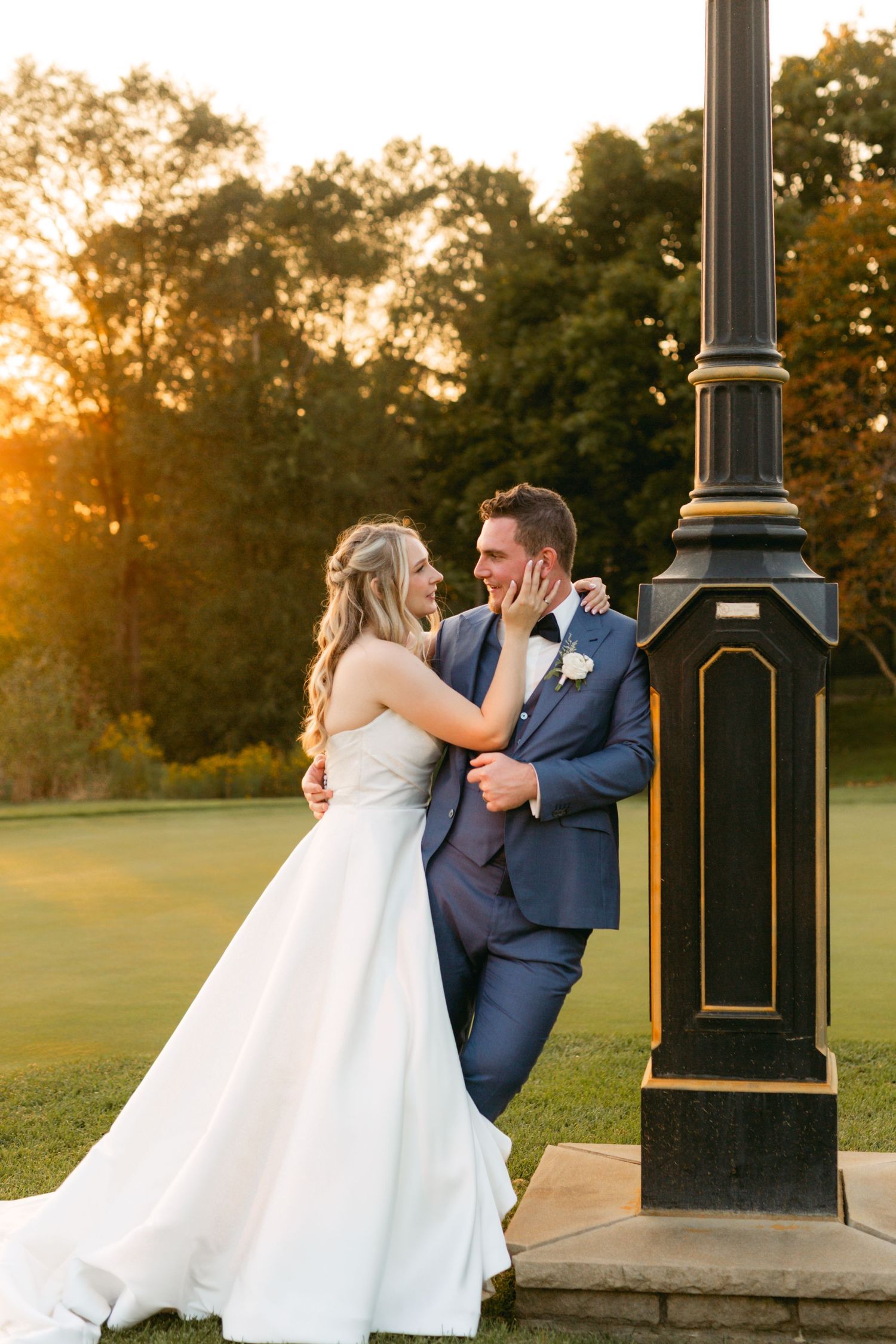 A romantic sunset wedding photoshoot features a couple posing playfully by a vintage clock post on a golf course.