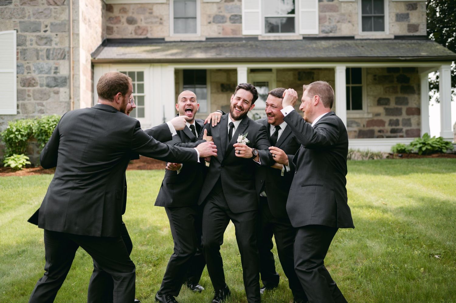 Groomsmen in black suits share laughs and celebrate on the lawn of a historic stone building.