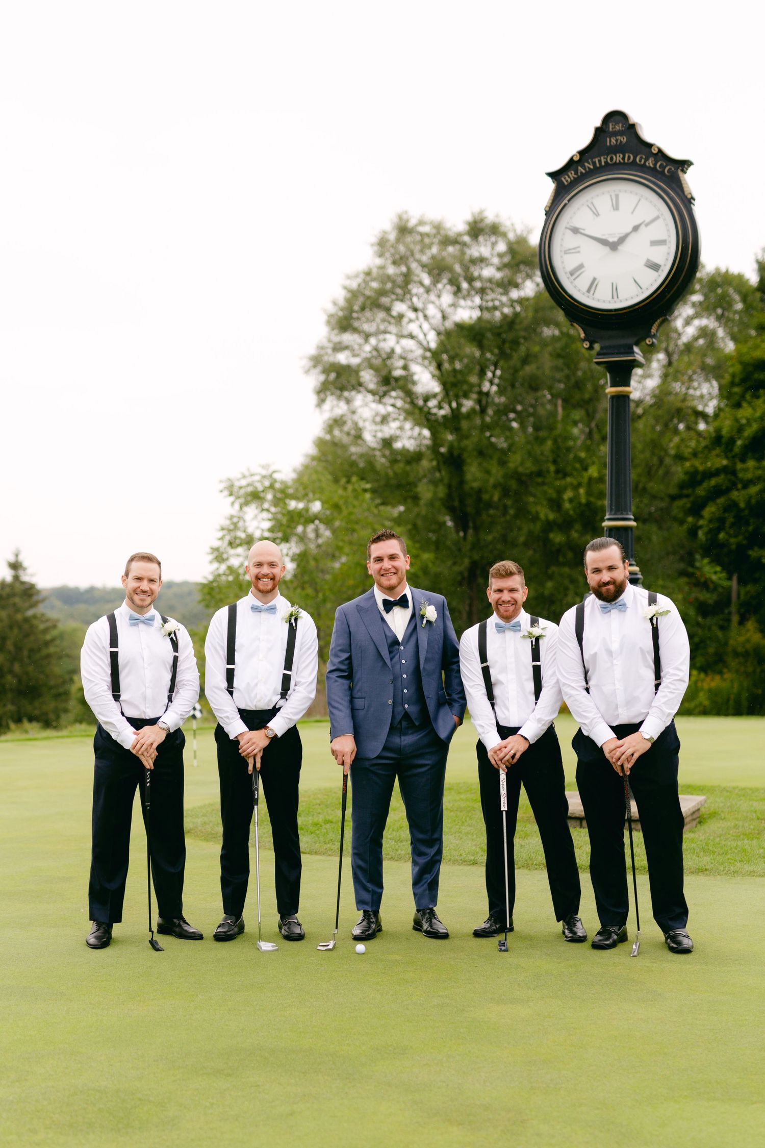 Wedding party in formal attire poses for group photo on golf course with decorative clock in background.