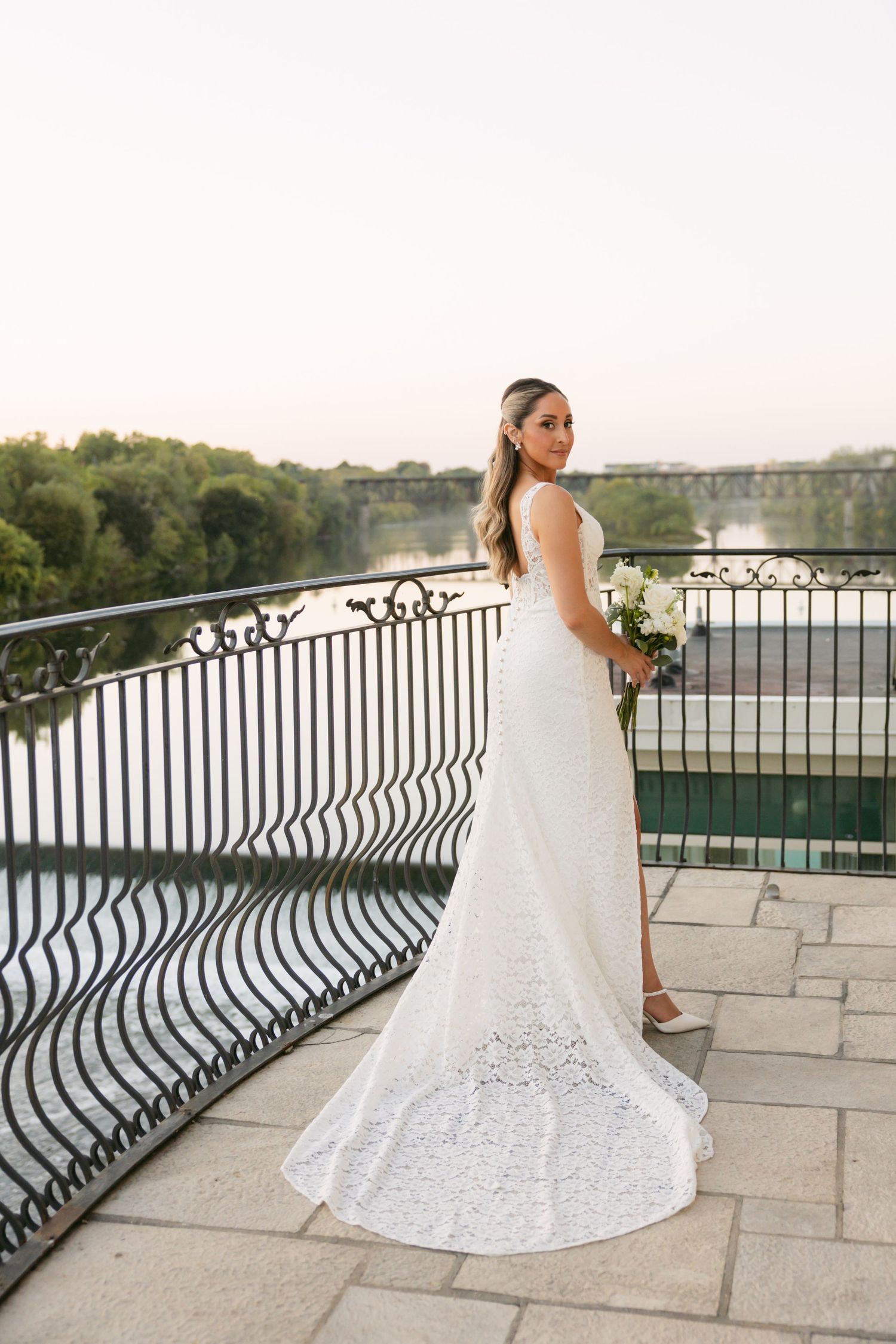 Bride in white lace gown poses by ornate railing with water view.