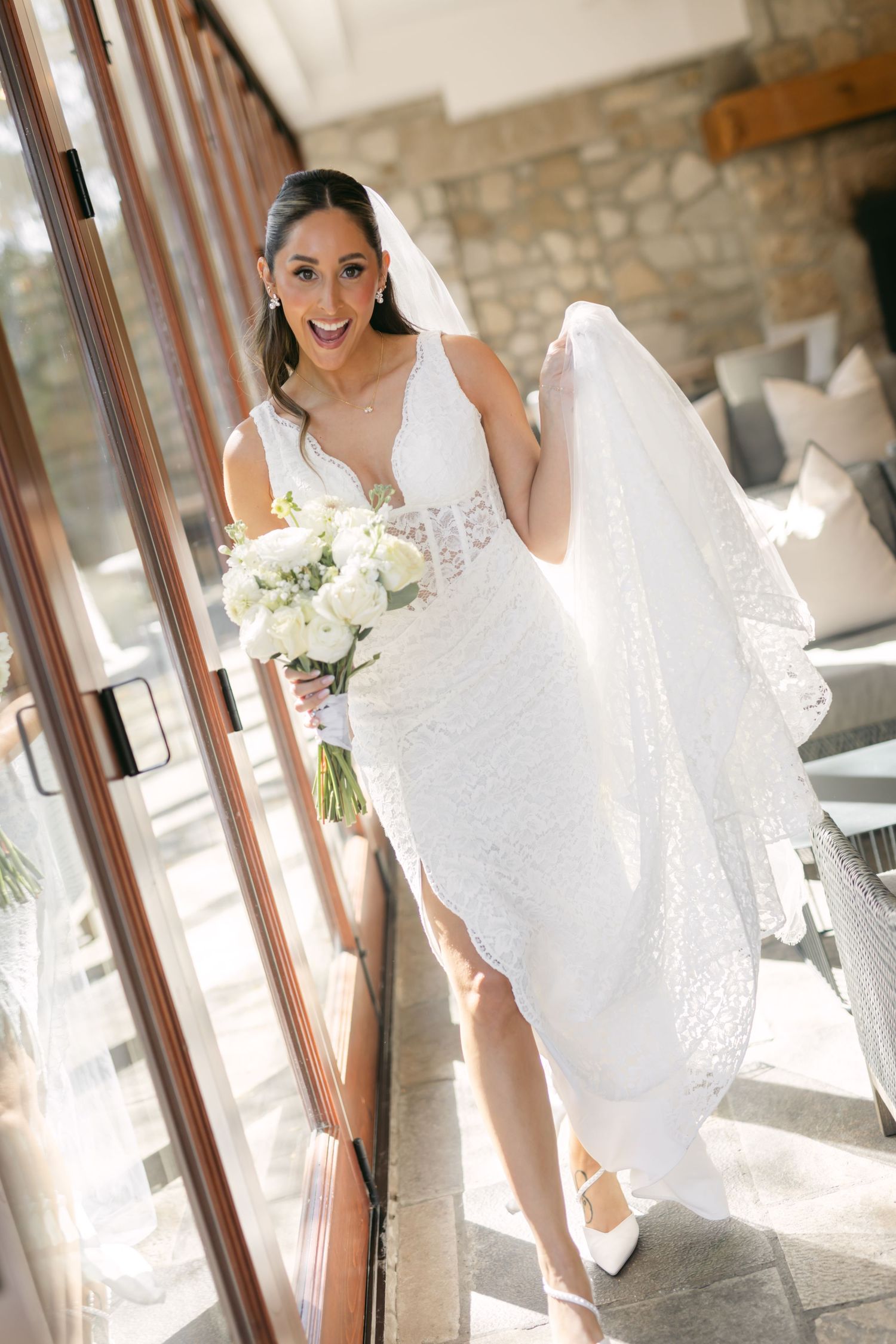 A bride in lace holds a white bouquet while posing near large windows.