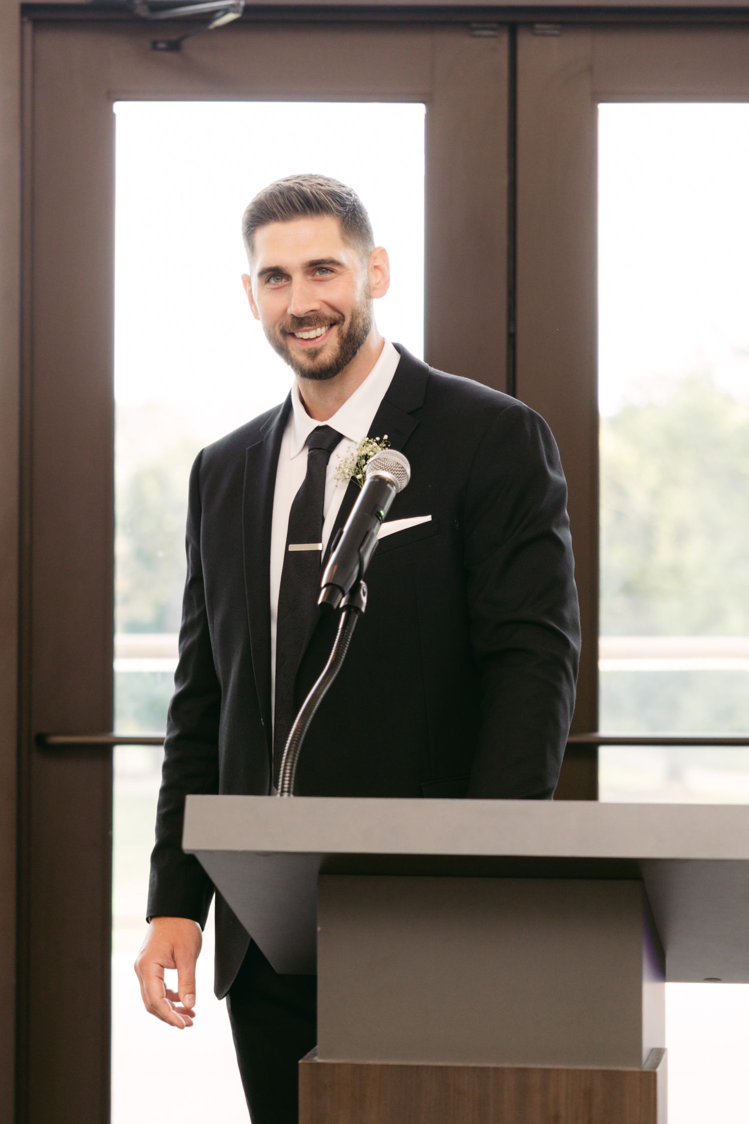 A groom in a black tuxedo poses against a modern interior backdrop with geometric shelving.