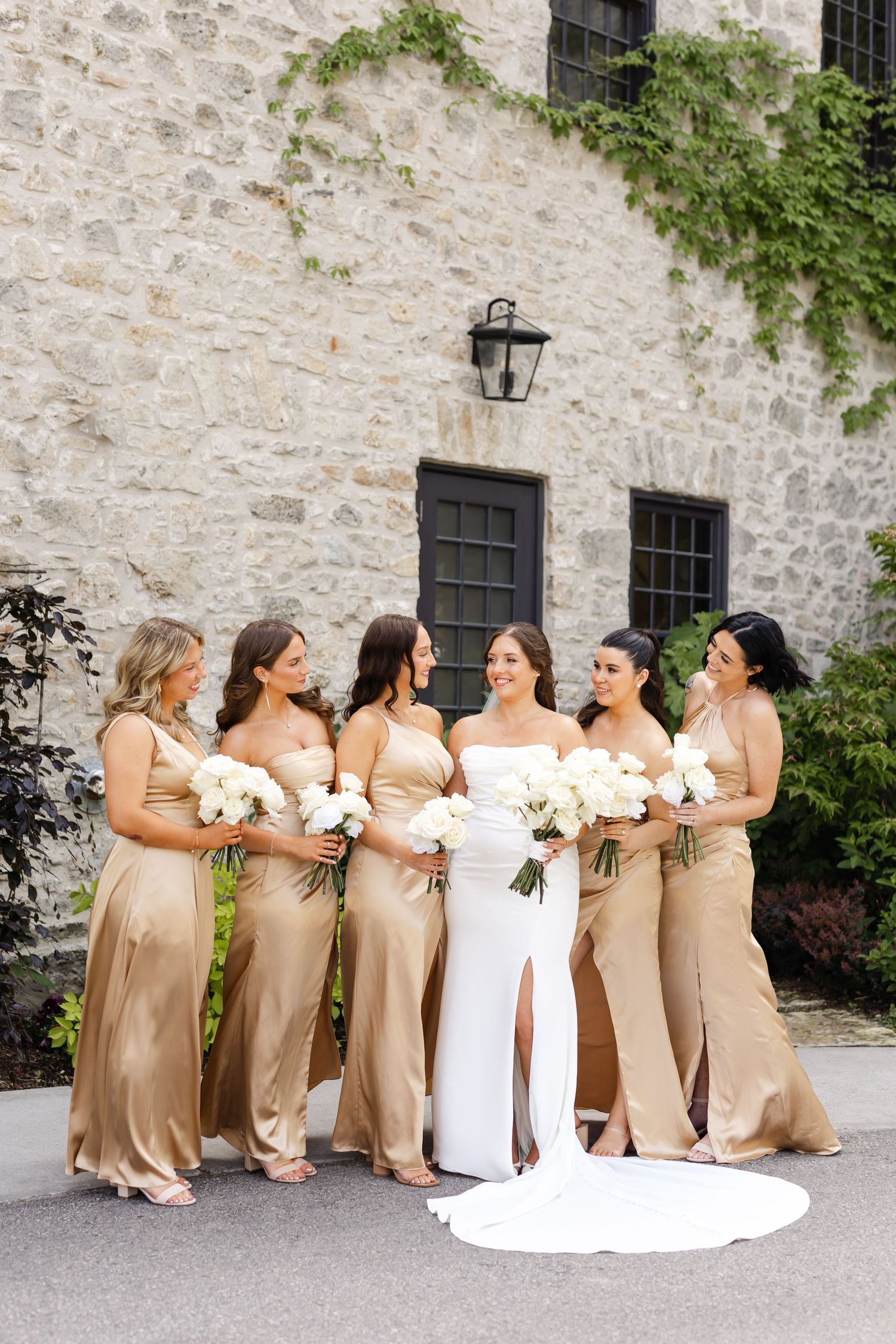 Bride in white gown with five bridesmaids in gold satin dresses holding white floral bouquets outside stone building.