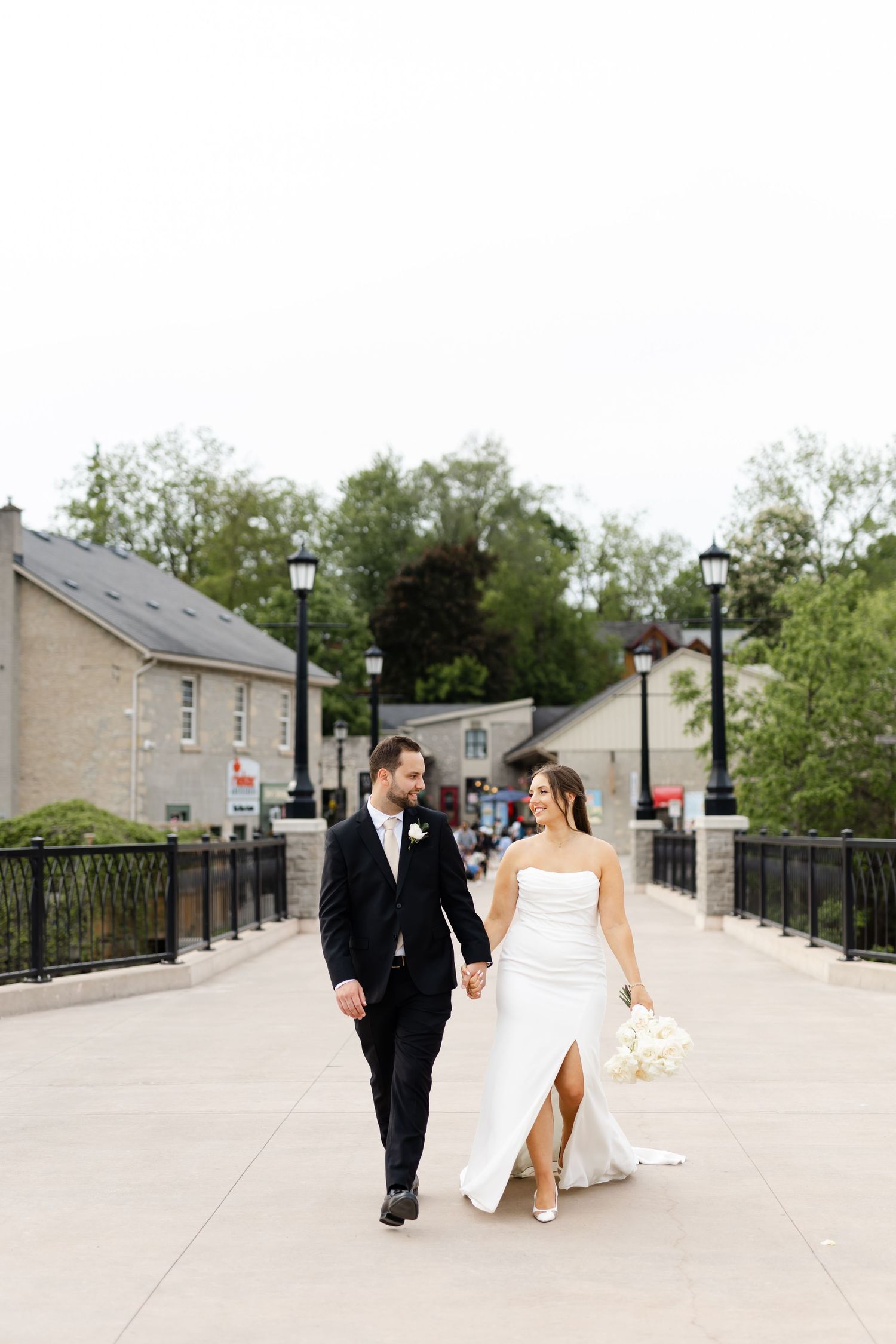 Bride and groom walk hand-in-hand on a bridge, bride in strapless white gown holding white bouquet.