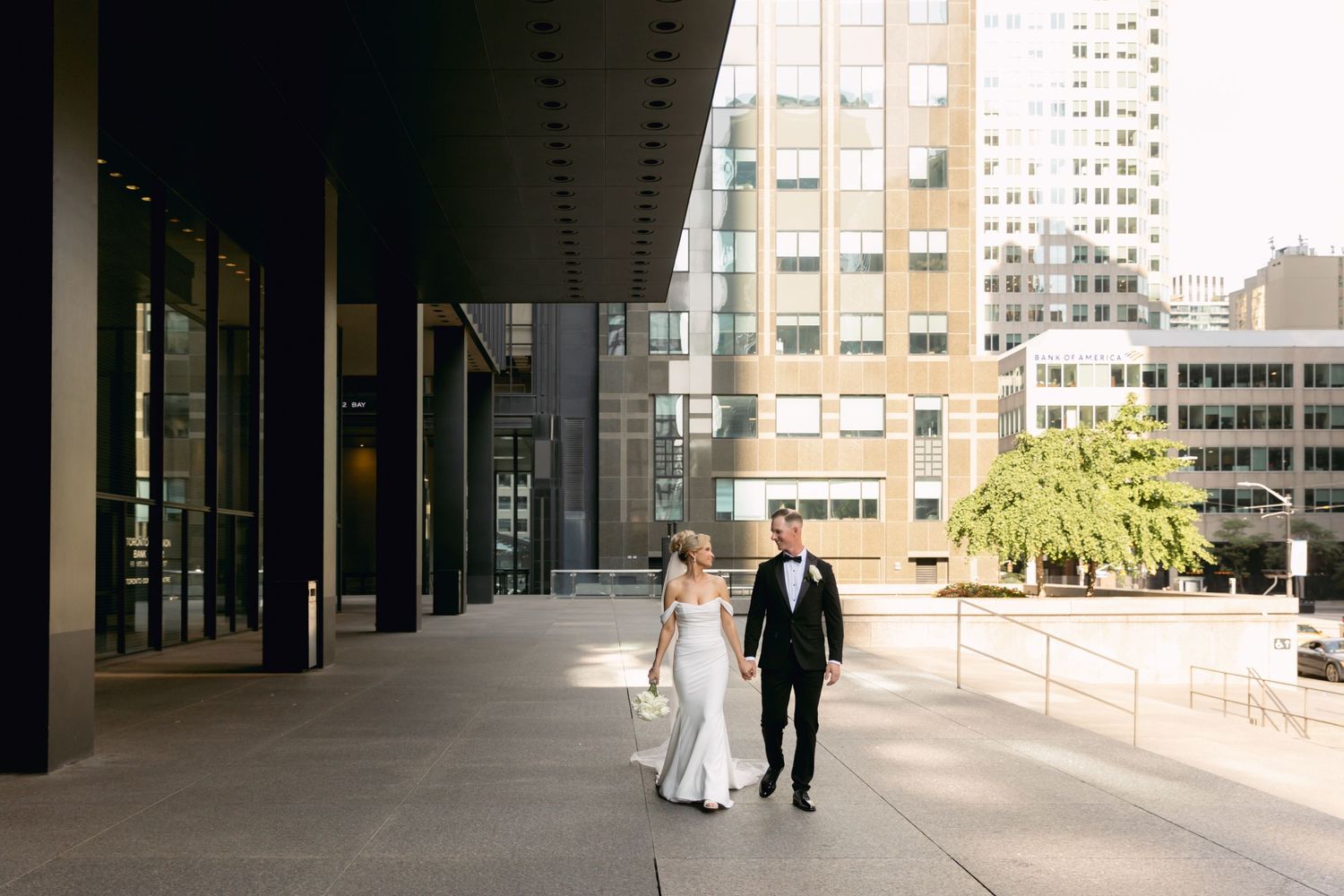 Bride in white gown and groom in black tux walk hand in hand along modern urban walkway.
