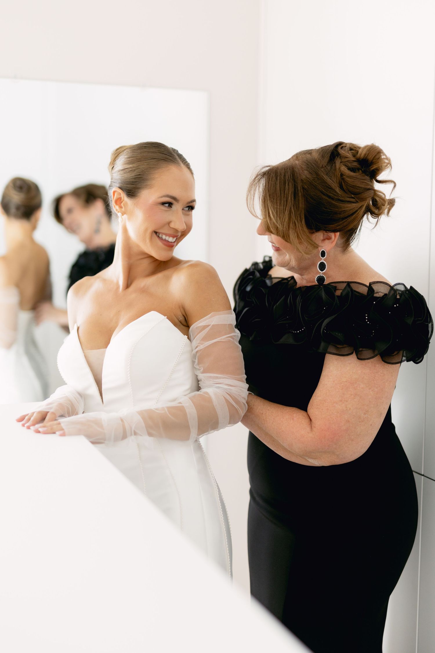 Smiling bride in strapless white gown with sheer gloves being helped by woman in black dress before mirror.
