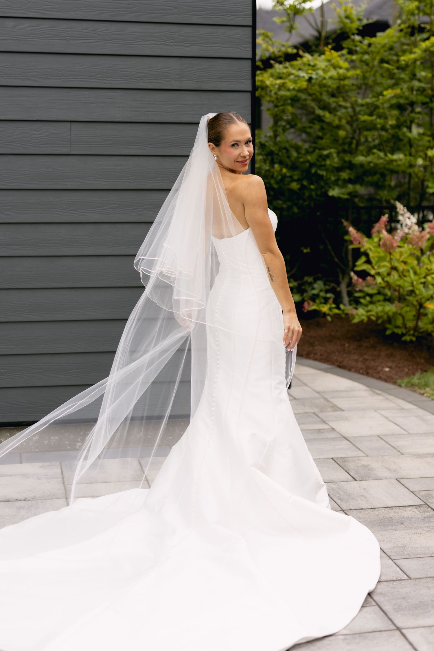 Bride in strapless white mermaid gown with flowing veil looks back over shoulder outdoors.