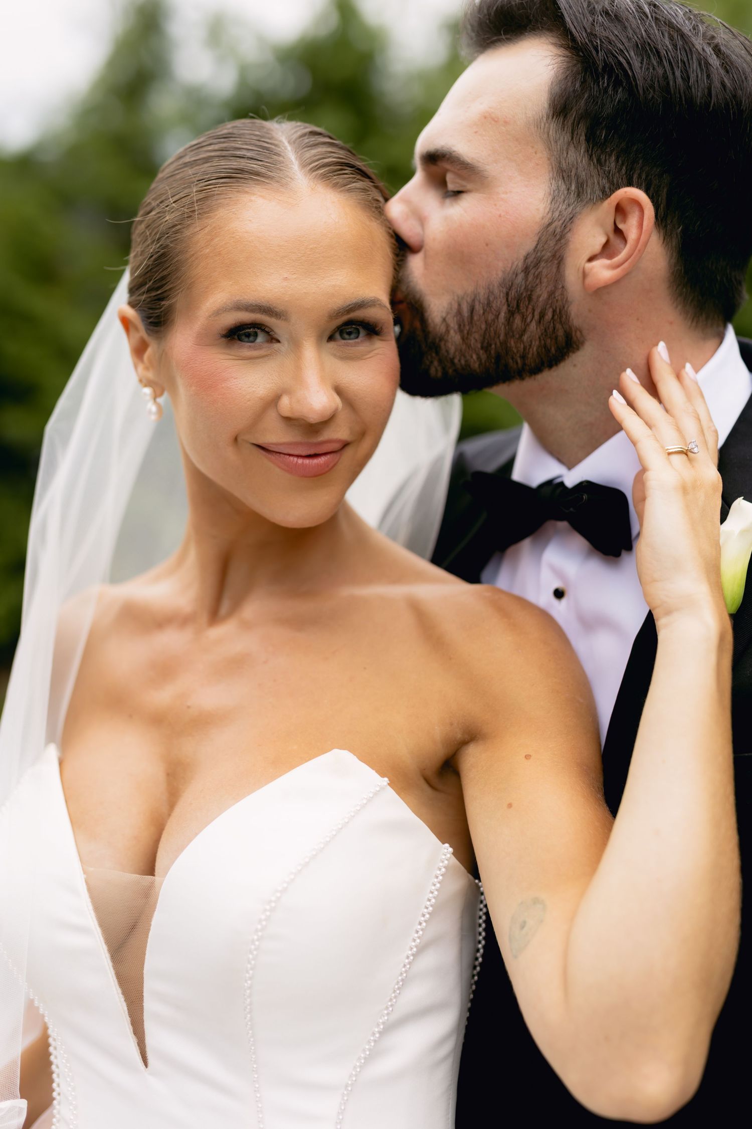 Groom tenderly kissing bride on cheek while she smiles at camera outdoors.