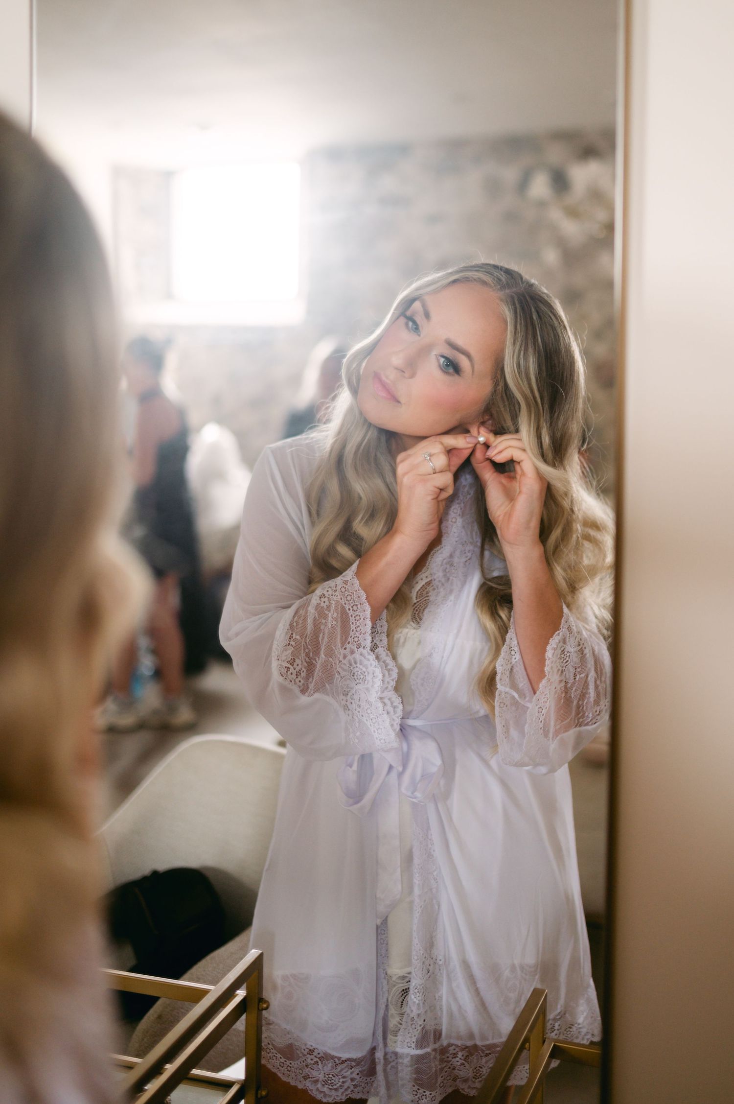 A person in a white lace robe gets ready in front of a mirror on their wedding morning.
