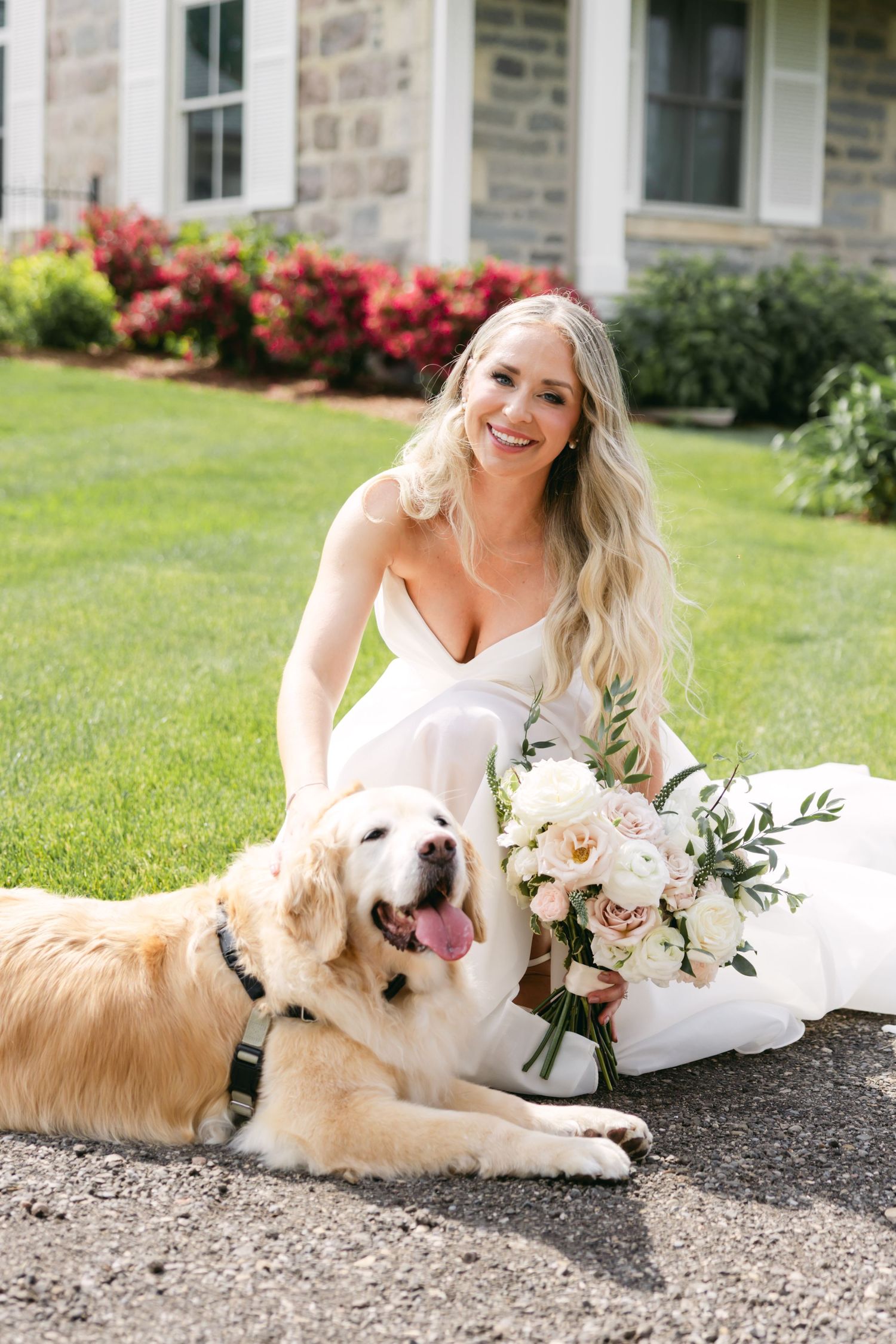 Bride in white wedding dress poses with golden retriever dog on gravel path.