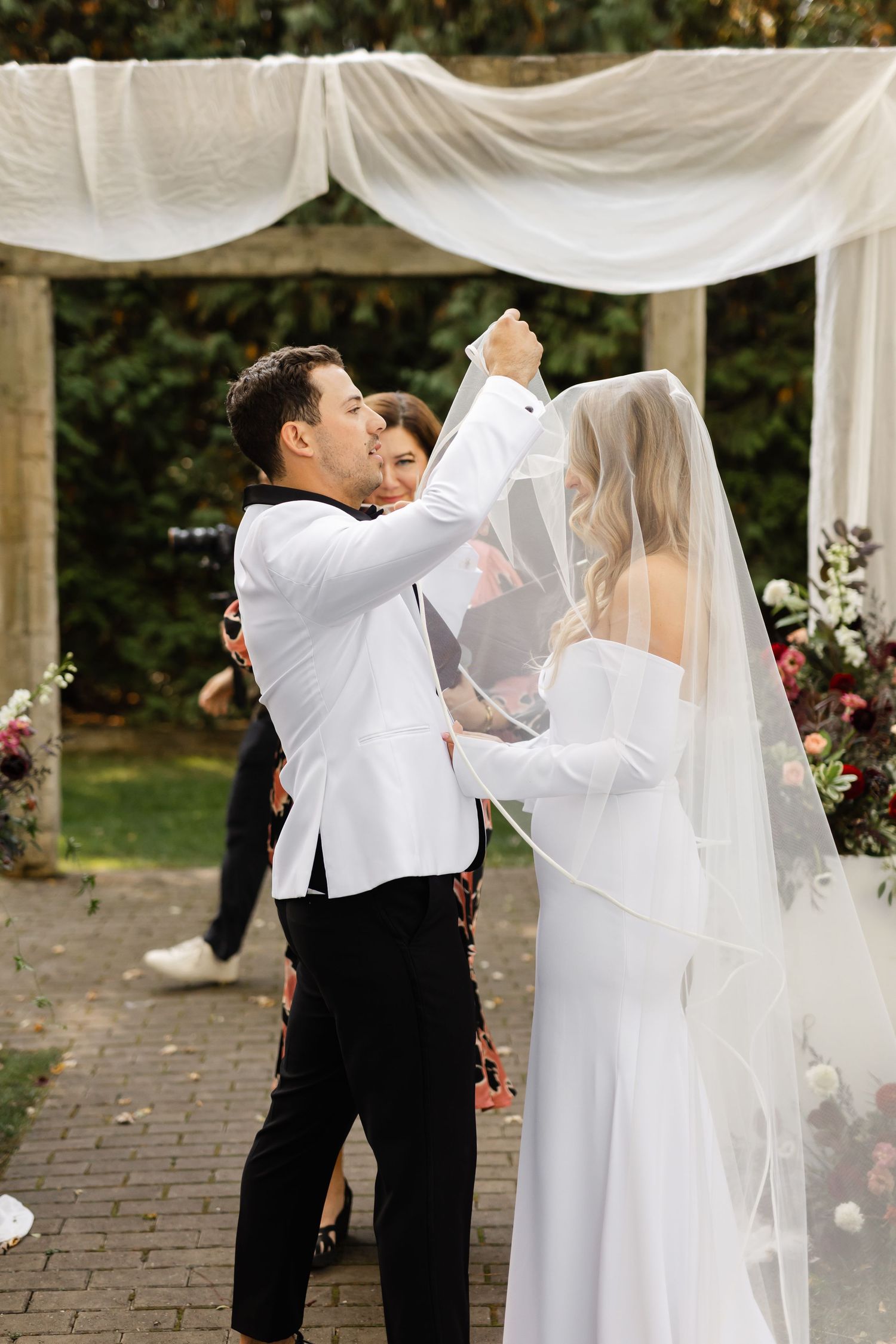 A wedding couple shares a playful moment with a flowing white veil in an outdoor ceremony setting.