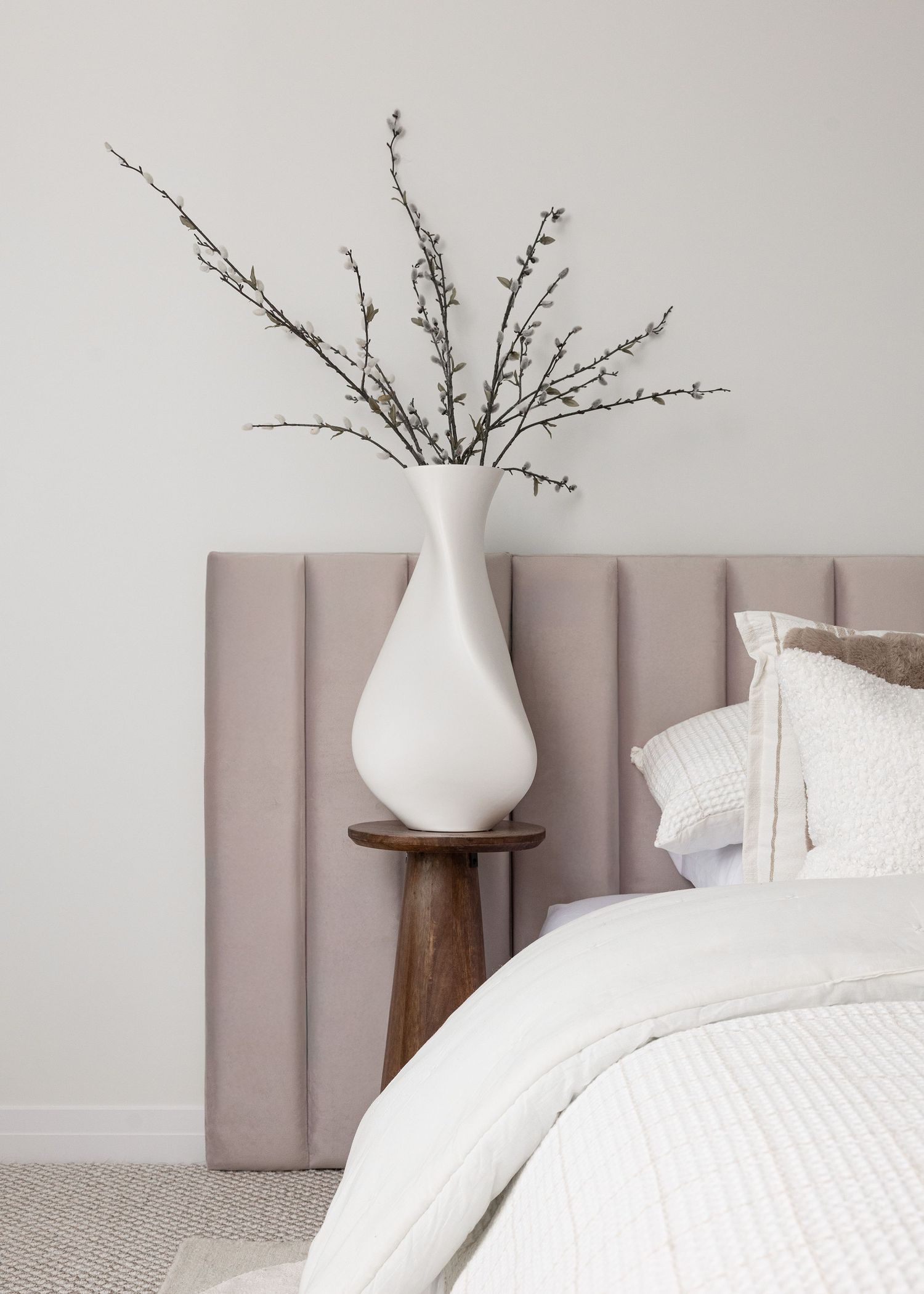 White ceramic vase with delicate branches displayed against a taupe channel-tufted headboard.