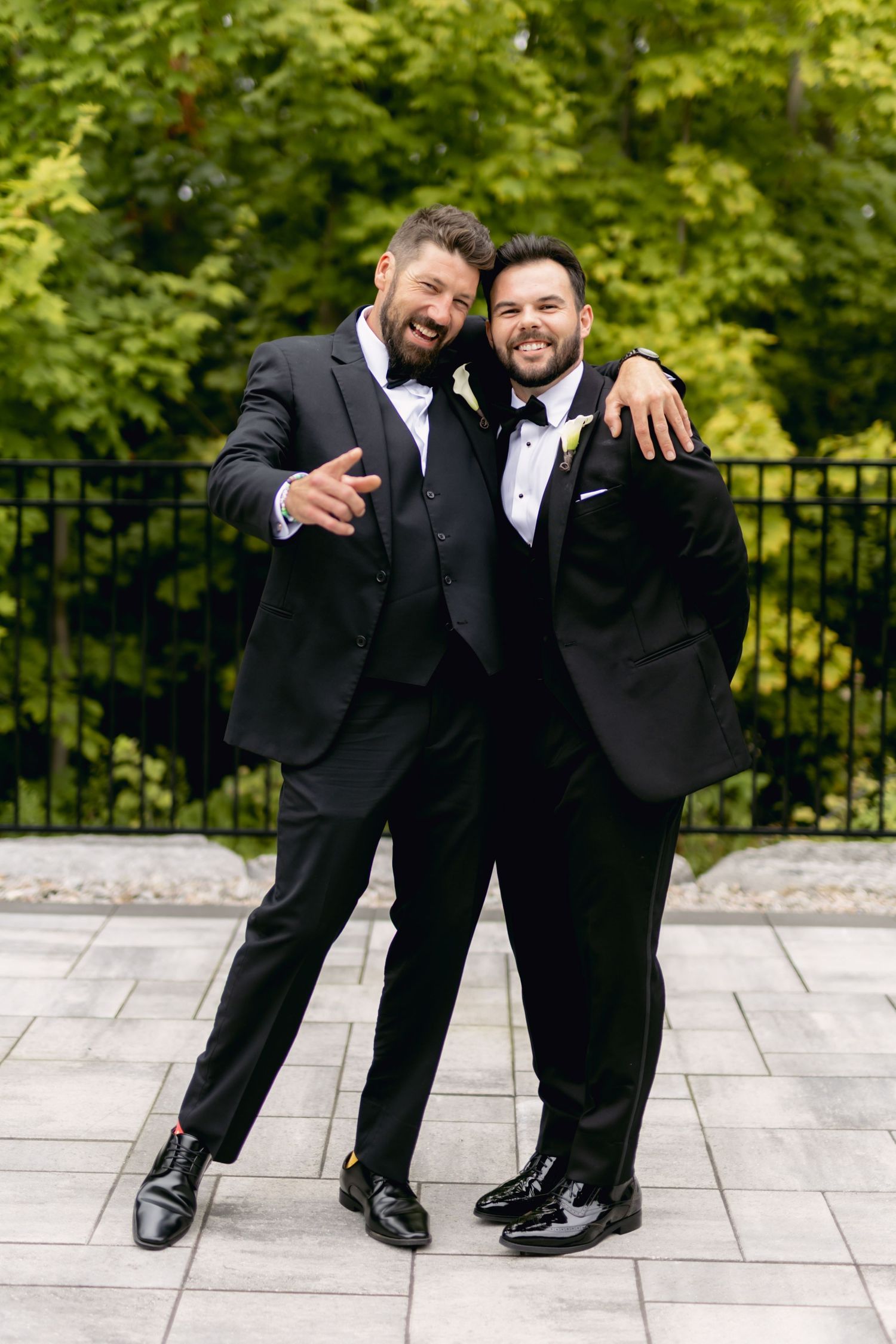 Two groomsmen in black tuxedos embrace and smile outdoors with lush green trees in background.