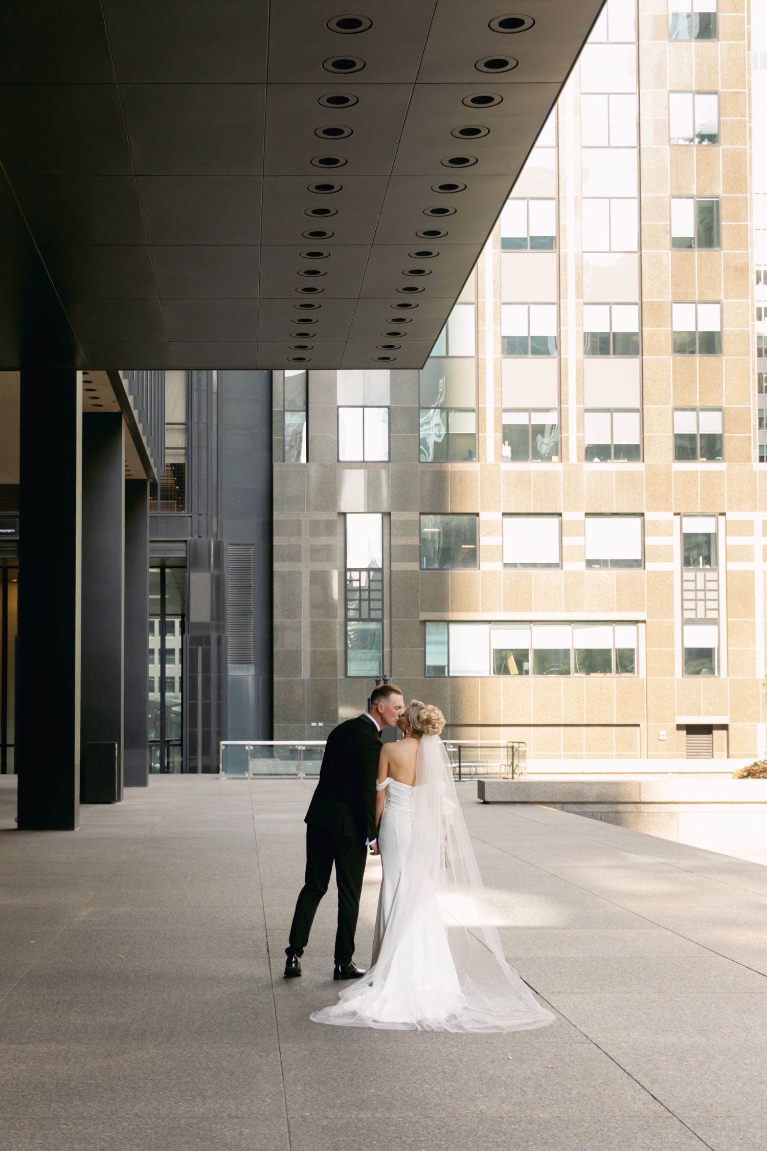 Bride and groom sharing romantic kiss outside modern glass skyscraper, bride in cathedral veil.