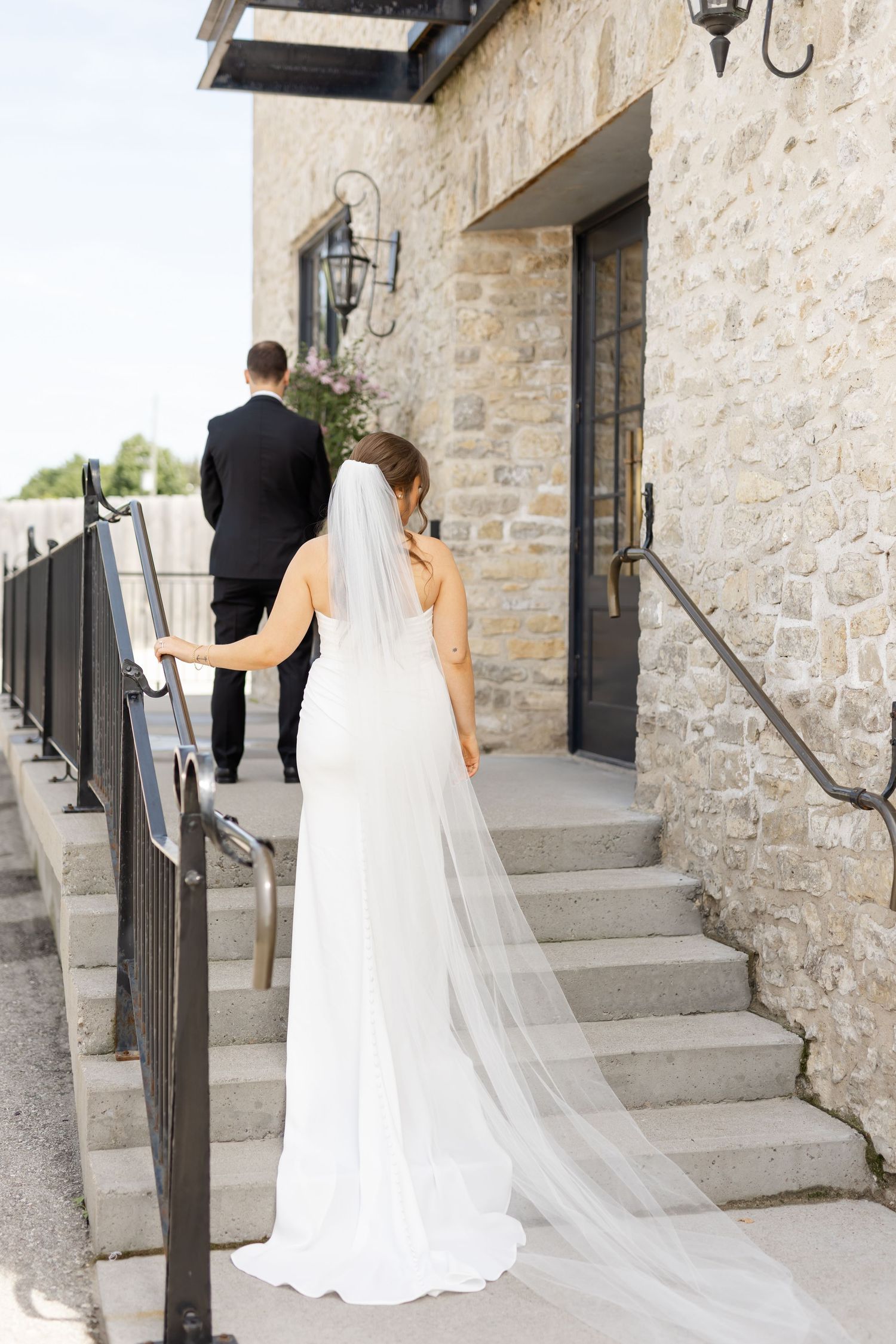Bride in white gown and veil walks up stone steps with groom in black suit at elegant venue.