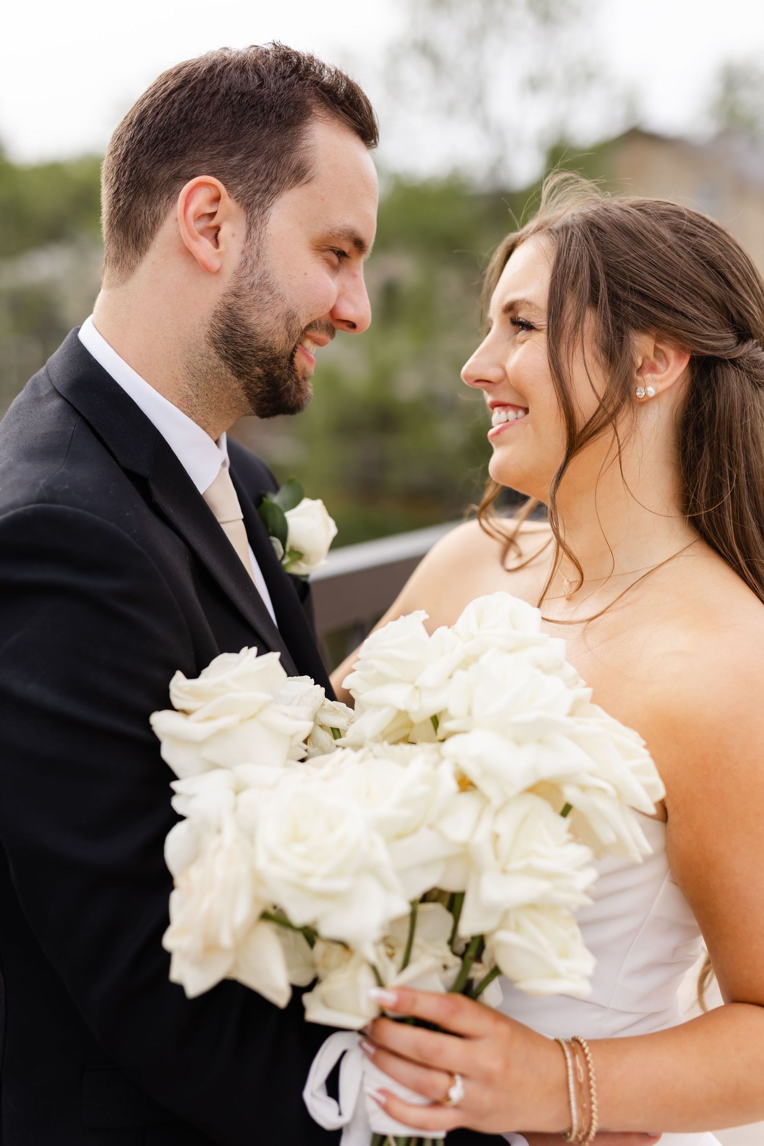 Groom gazes lovingly at smiling bride as she holds a lush bouquet of white roses on wedding day.