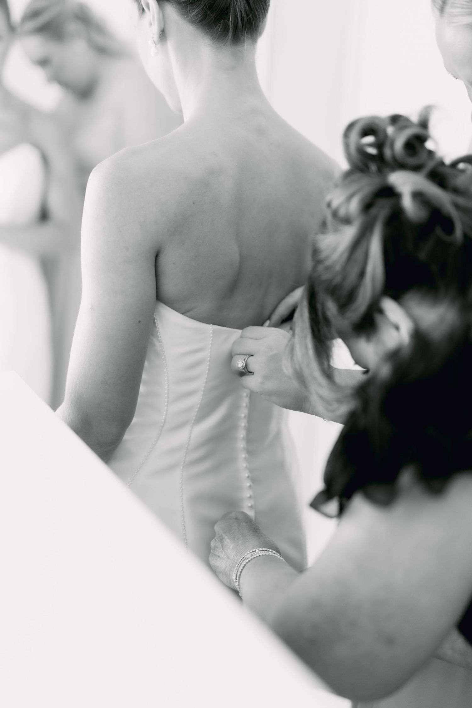 Black and white photo of bridesmaid buttoning back of brides fitted wedding gown with pearl buttons.