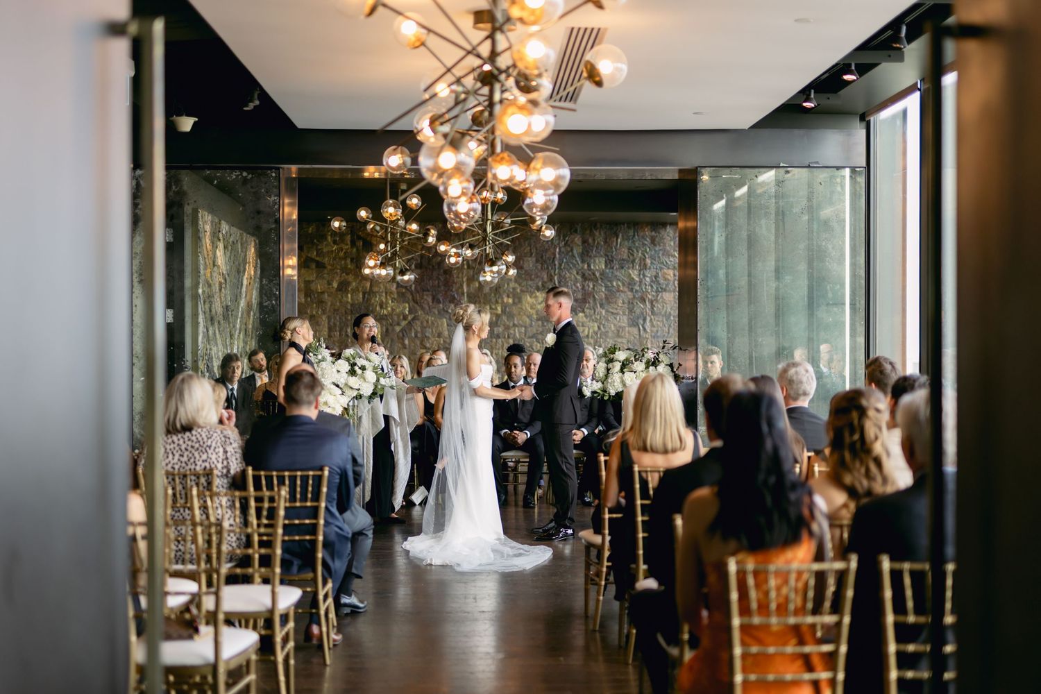 Wide shot of intimate wedding ceremony with bride and groom at altar surrounded by seated guests in modern venue.